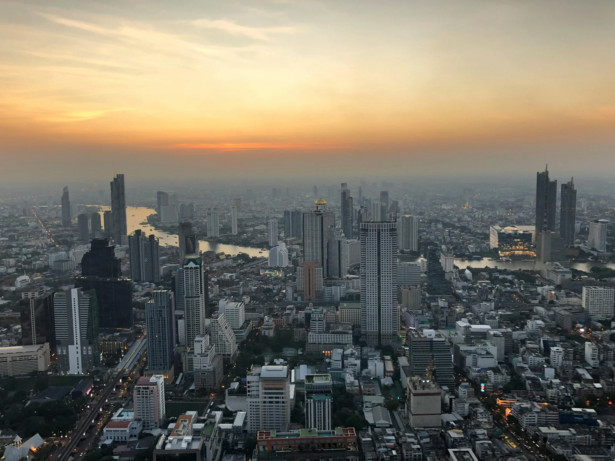 A city skyline at sunset with tall skyscrapers, a river, and a sky with orange, pink, and blue hues.