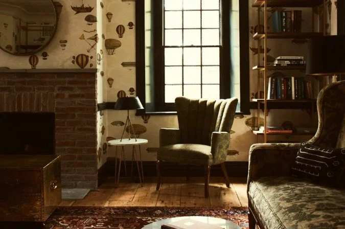 An interior living room with a brick fireplace, a round mirror, a window, an armchair, a small side table with a lamp, a bookshelf with books, a patterned sofa, and a vintage-style rug.