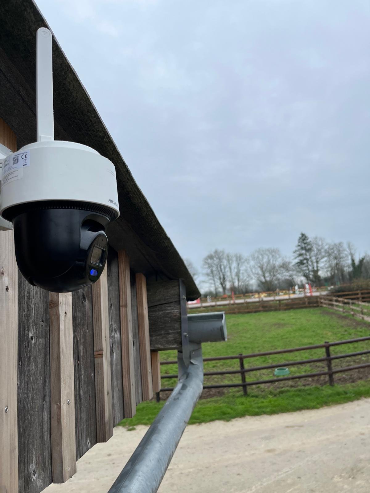 Caméra de surveillance installée à l'extérieur sur un bâtiment en bois, avec un paysage de champ et d'arbres en arrière-plan, sous un ciel nuageux.