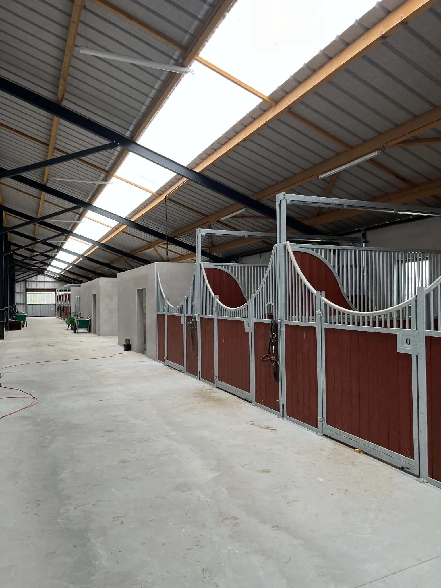Intérieur d'une écurie avec des boxes pour chevaux avec portes en bois et en métal, le toit en tôle et une grande ouverture permettant la lumière naturelle.