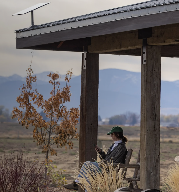 Une femme assise sur un banc en bois sous un abri en bois, regardant son téléphone, avec un paysage de champs et une antenne Starlink sur le toit du bâtiment.
