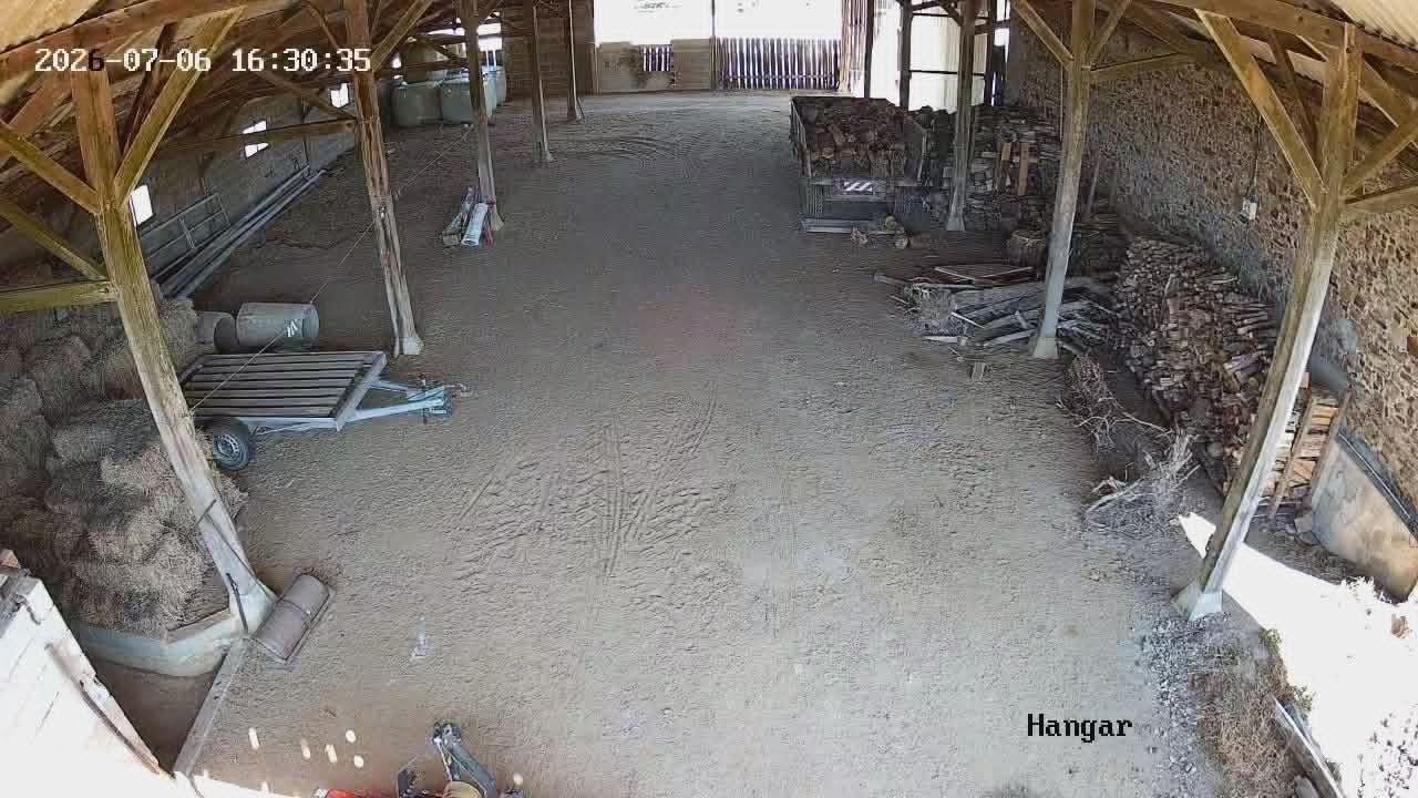 Intérieur d'un hangar en construction ou rangement, avec un sol en terre, des piles de bois et des outils empilés sur le côté, structure en bois et murs en pierre visible.