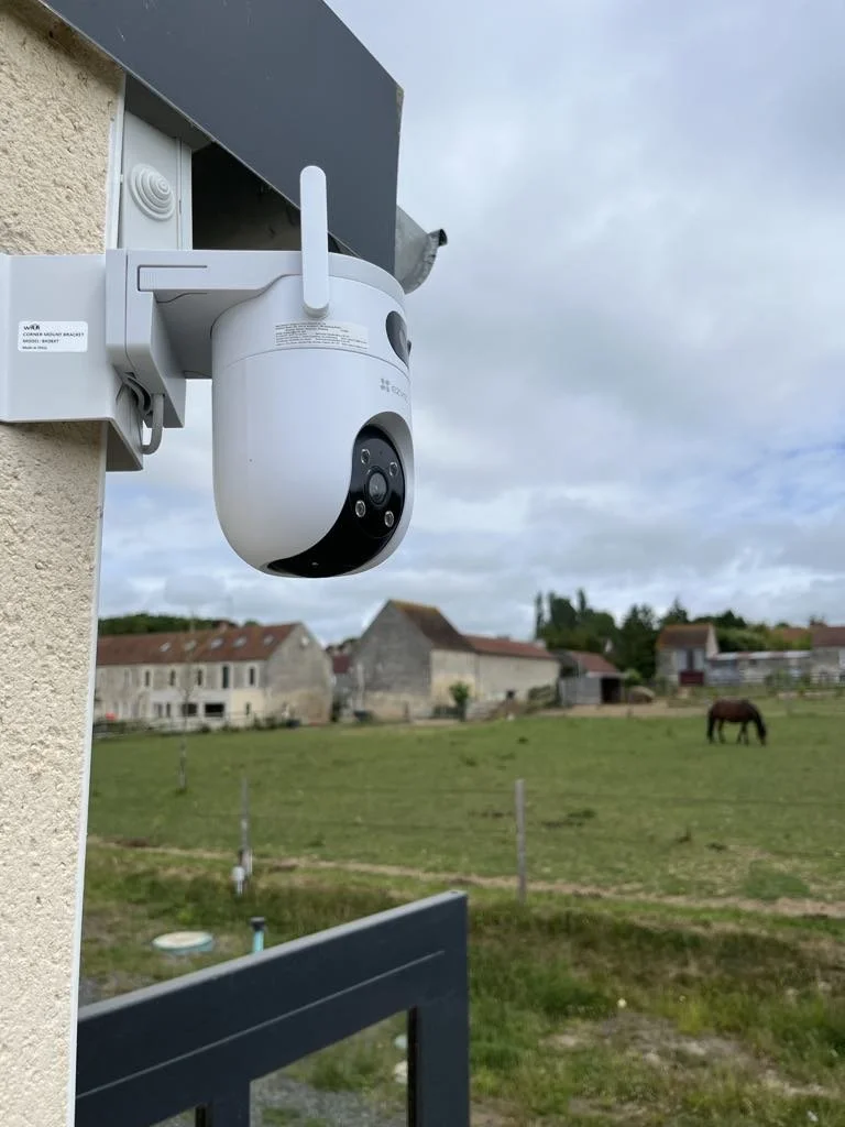 Caméra de surveillance installée à l'extérieur près d'un bâtiment avec un paysage rural en arrière-plan, comprenant des maisons, un cheval dans un pré et un ciel nuageux.