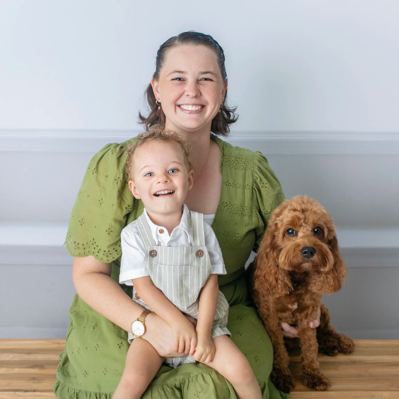 A smiling woman wearing a green dress sitting on a wooden floor with a young boy sitting on her lap, a brown curly-haired dog sitting beside them, all posing for the photo against a plain light-colored wall.