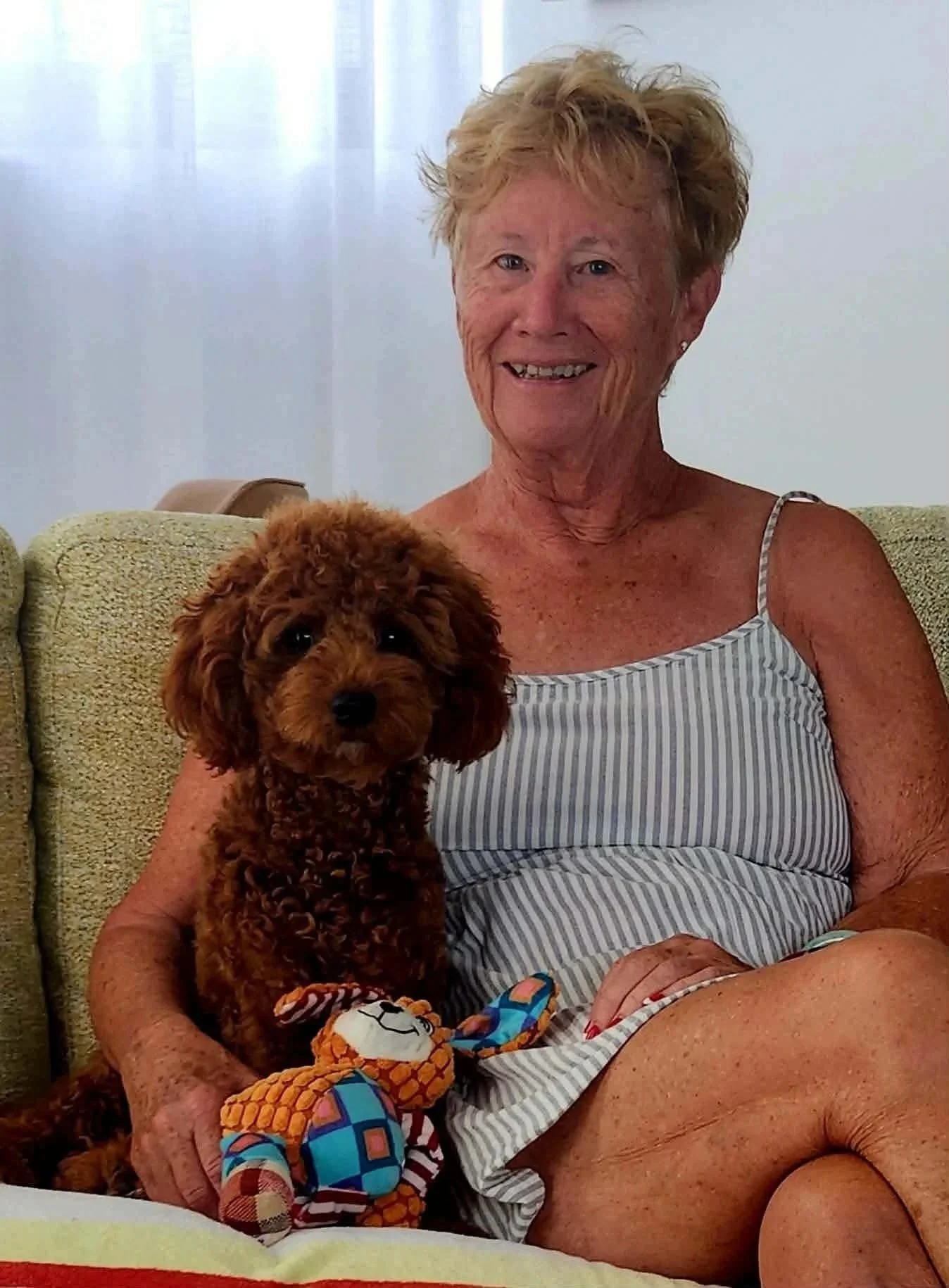 An elderly woman with short, curly blonde hair smiling while sitting on a green couch with a small brown poodle in her lap, wearing colorful socks and a striped dress.