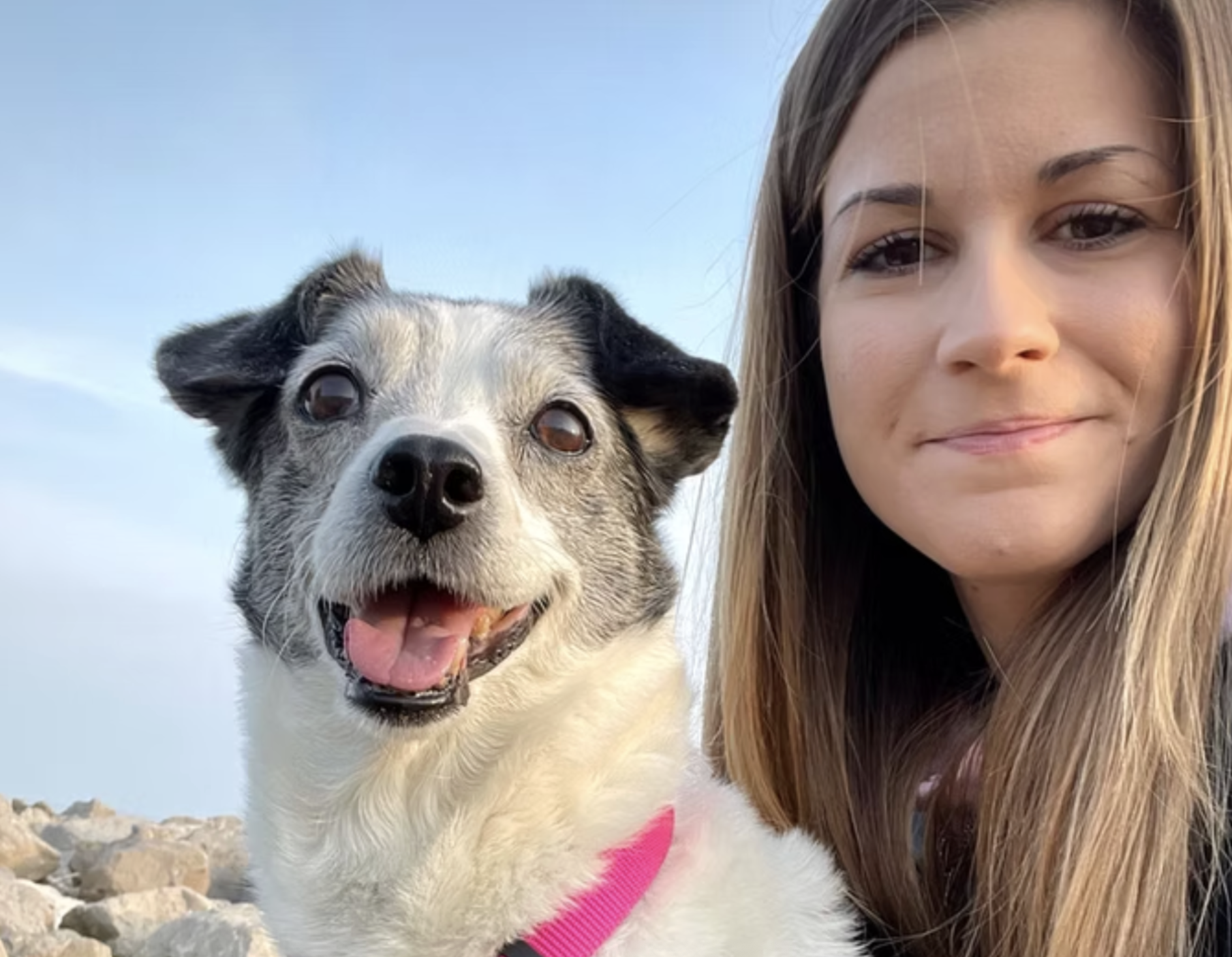 Una donna con lunga chioma castana e un cane con pelo bianco e nero sorridenti, con sfondo di cielo azzurro e rocce.