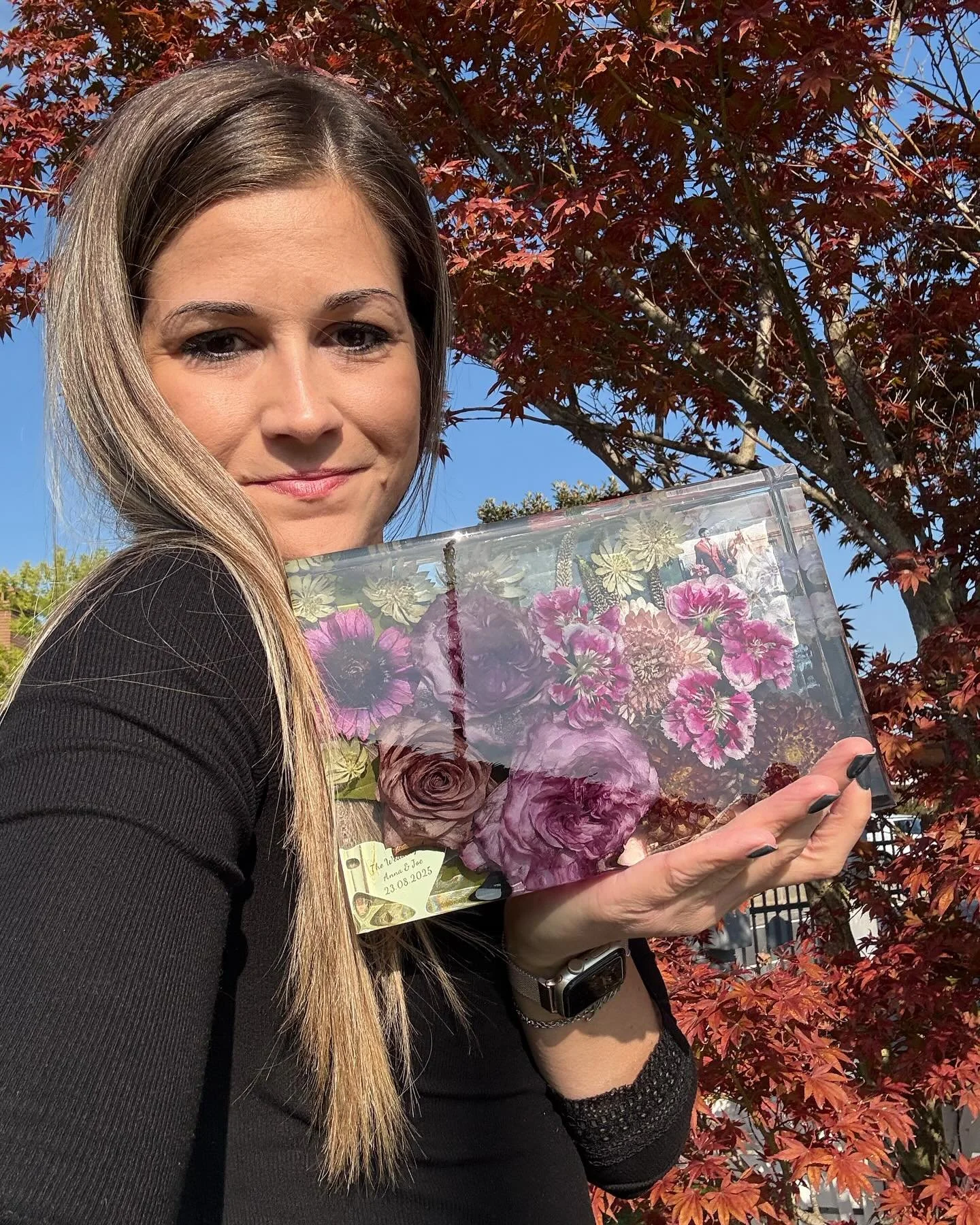 Una donna con capelli lunghi e chiari tiene un una creazione in resina di fiori rosa e viola, con uno sfondo di foglie di un albero rosso e un cielo blu chiaro.