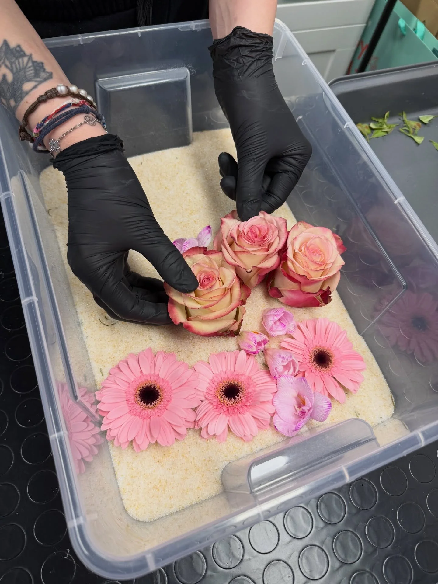 Due mani con guanti neri sistemano rose e fiori in uno stampo per resina.