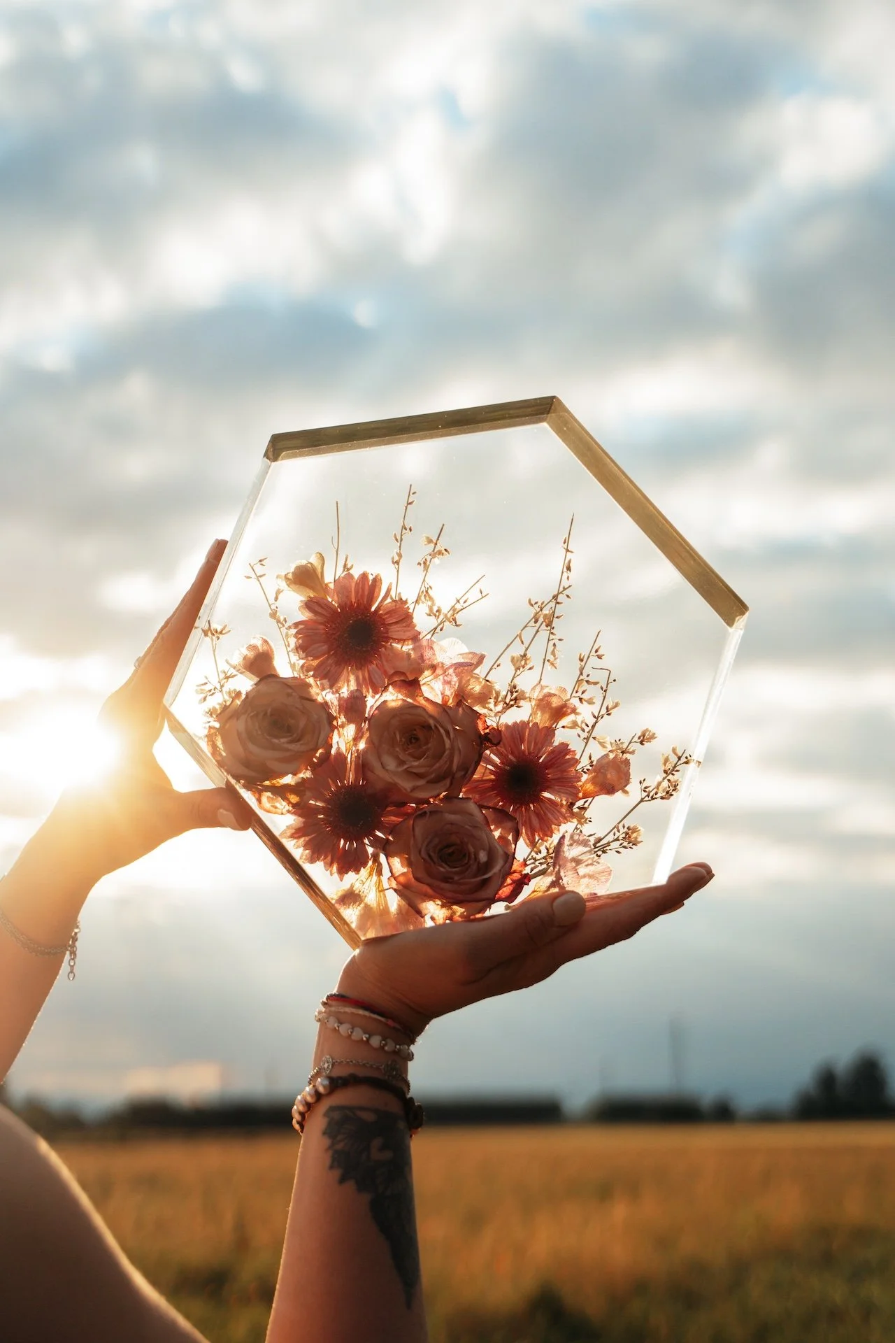 Una persona tiene in mano un oggetto decorativo di resina con fiori colorati, con il sole dietro e un cielo nuvoloso.