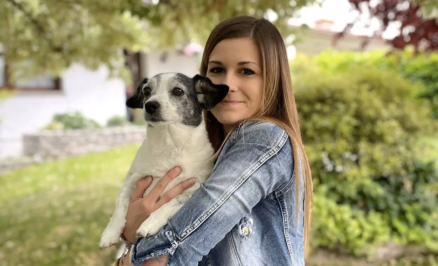 Una donna con capelli castani, vestita con una giacca di jeans, tiene un cane bianco e nero in braccio mentre sorride e si trova all'aperto, con alberi e case sullo sfondo.