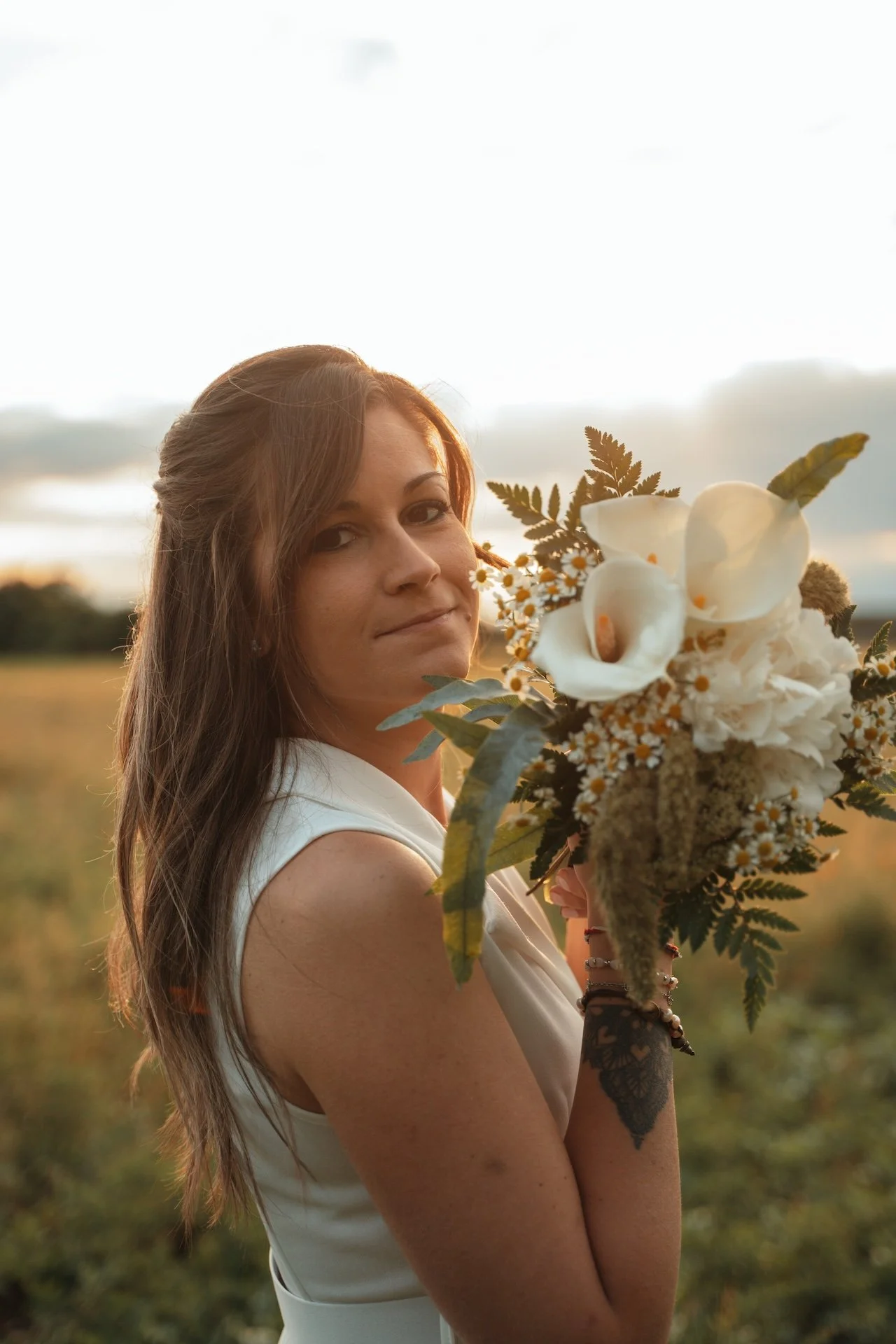 Una donna con i capelli lunghi e castani, indossa un vestito bianco, tiene un bouquet di fiori bianchi e verdi, si trova in un campo durante il tramonto.