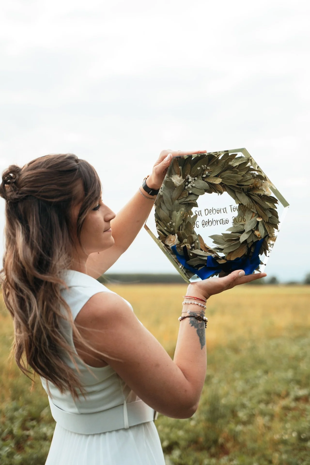 Una donna con capelli castani mossi, indossa un vestito bianco senza maniche e tiene Una creazione di resina di una corona di laurea con un nastro blu, con un campo aperto sullo sfondo.