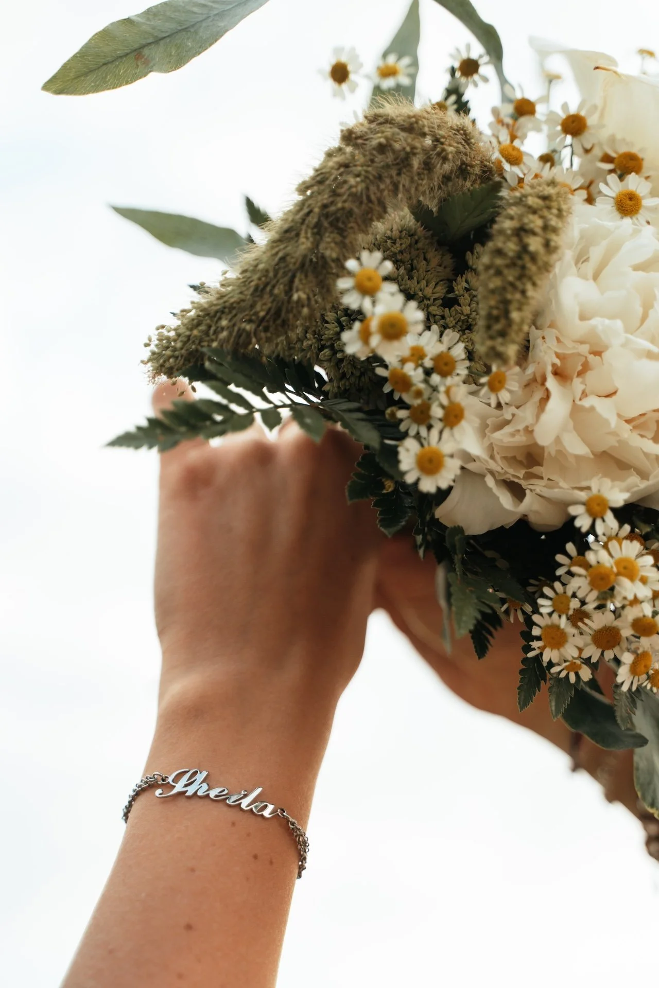 Mano con braccialetto chiaramente visibile, che indossa un anello con il nome "Gheila", tiene un bouquet di fiori bianchi e gialli con foglie verdi, sfondo bianco.