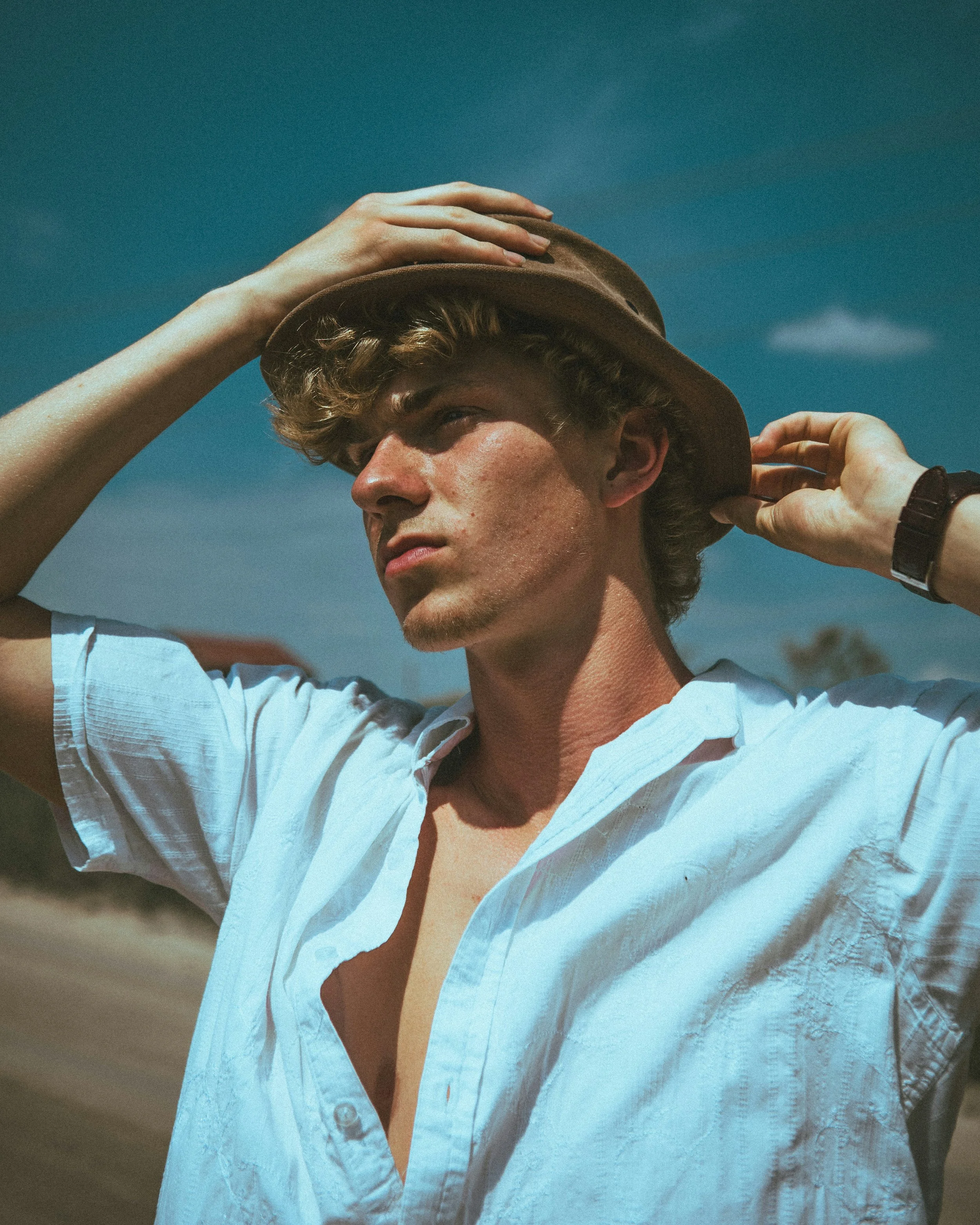 A young man outdoors wearing a light blue button-up shirt and a brown hat, touching the brim with one hand, against a blue sky background.