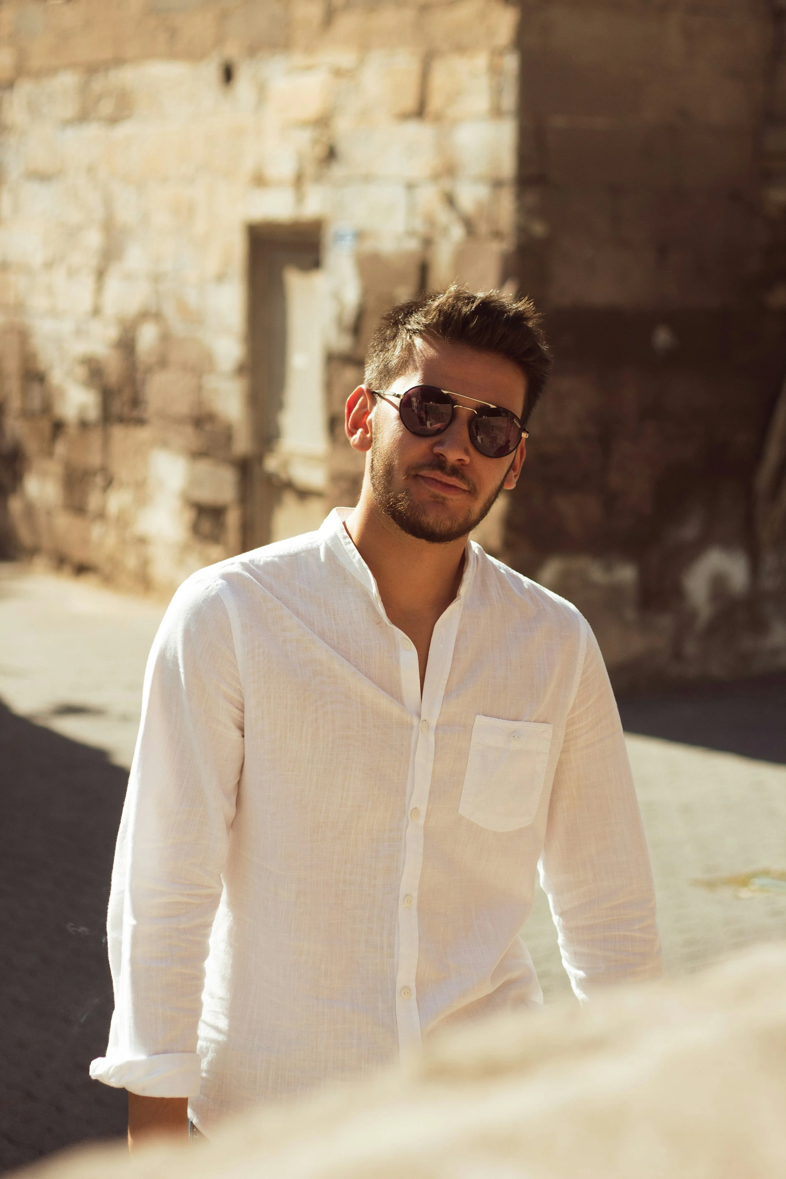 A young man wearing a white shirt and sunglasses standing outdoors with a blurred stone building background.