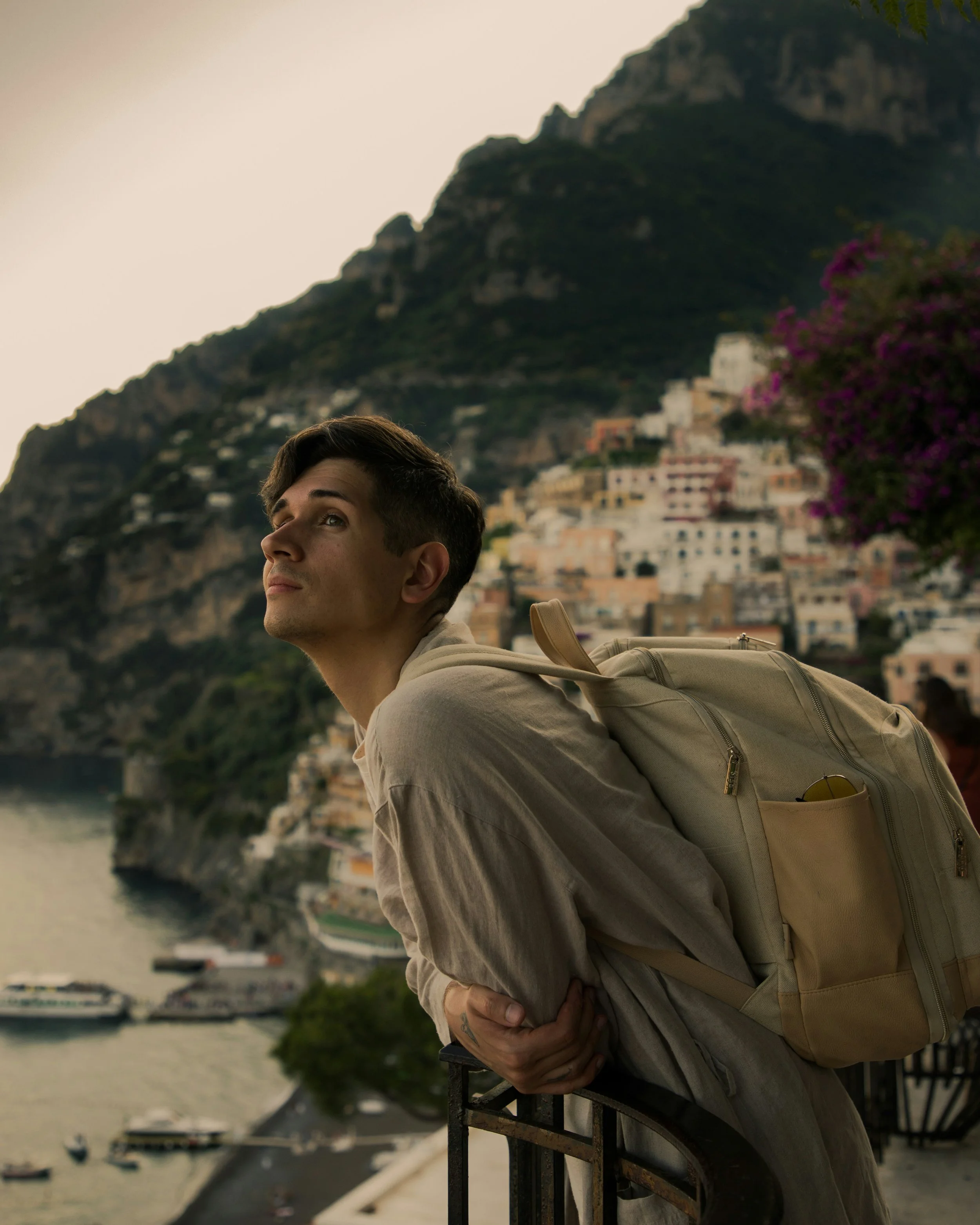 A young man with short brown hair, wearing a gray hoodie and beige backpack, leaning on a railing with a scenic hillside town with colorful buildings and a mountain in the background.