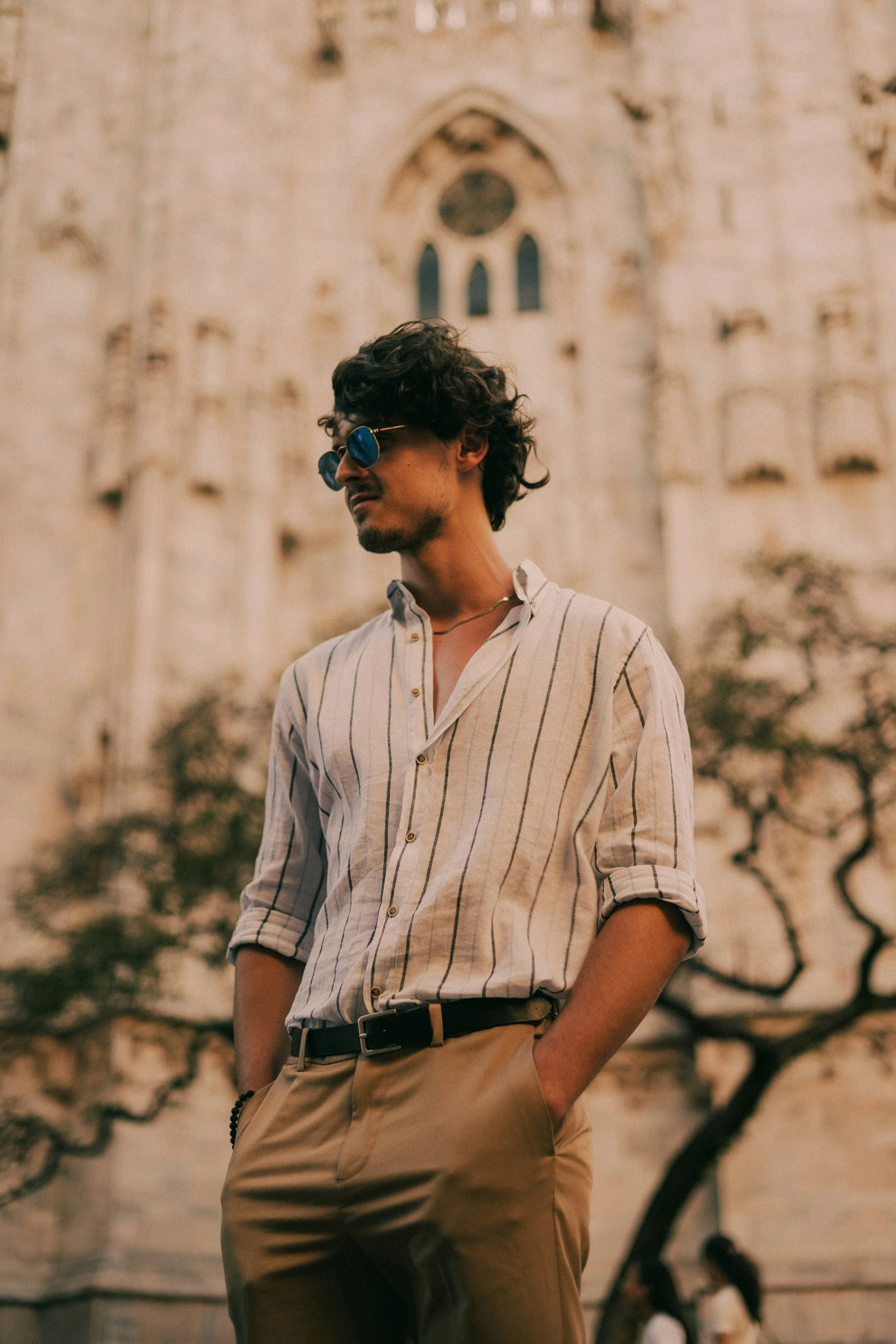 A man with curly hair and sunglasses standing outdoors against a historic stone building with an arched window in the background. He is wearing a beige and white striped shirt with the sleeves rolled up and beige pants with a black belt.