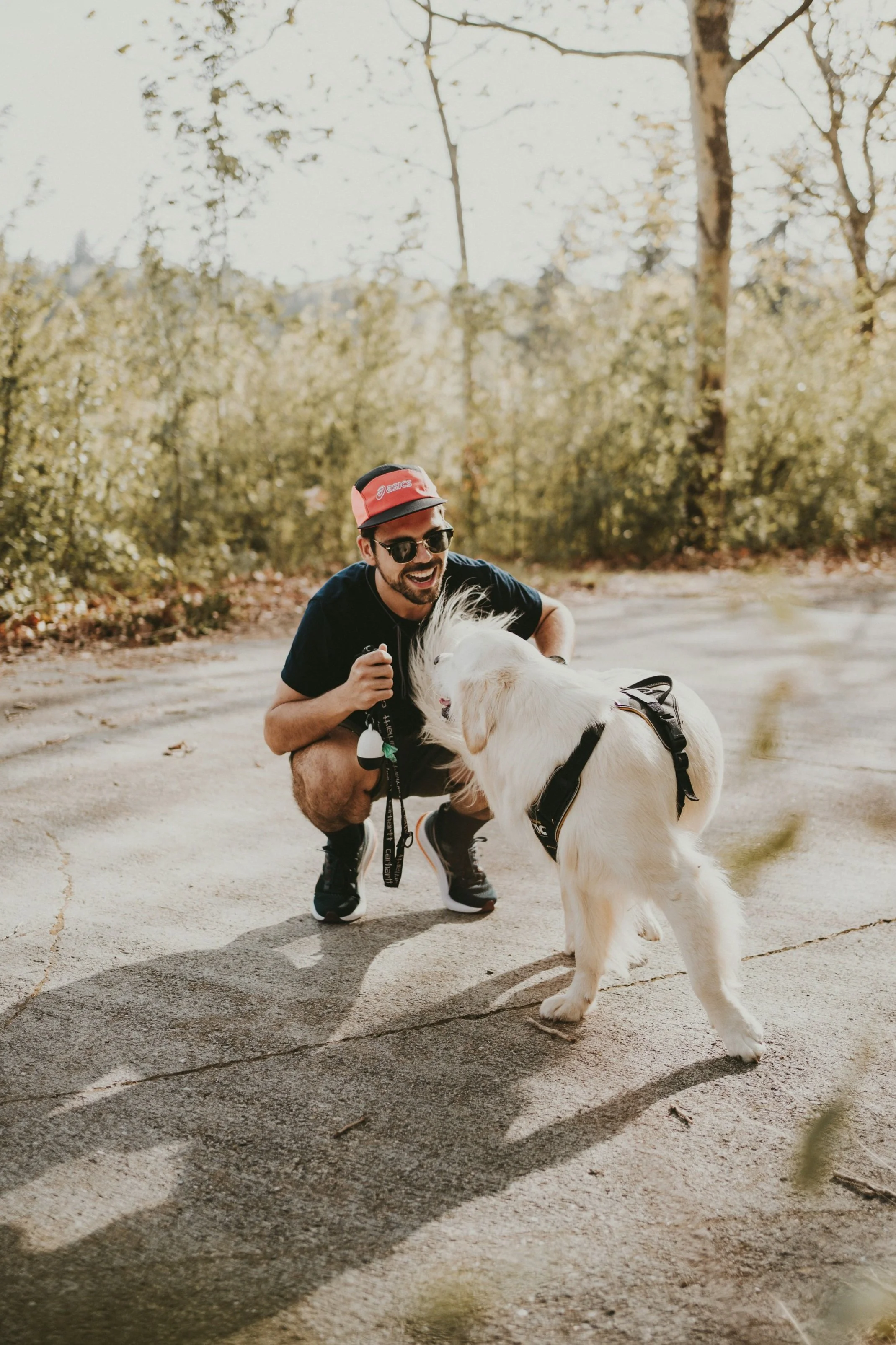 A man crouching and smiling at a white retriever puppy outdoors on a paved path surrounded by trees.