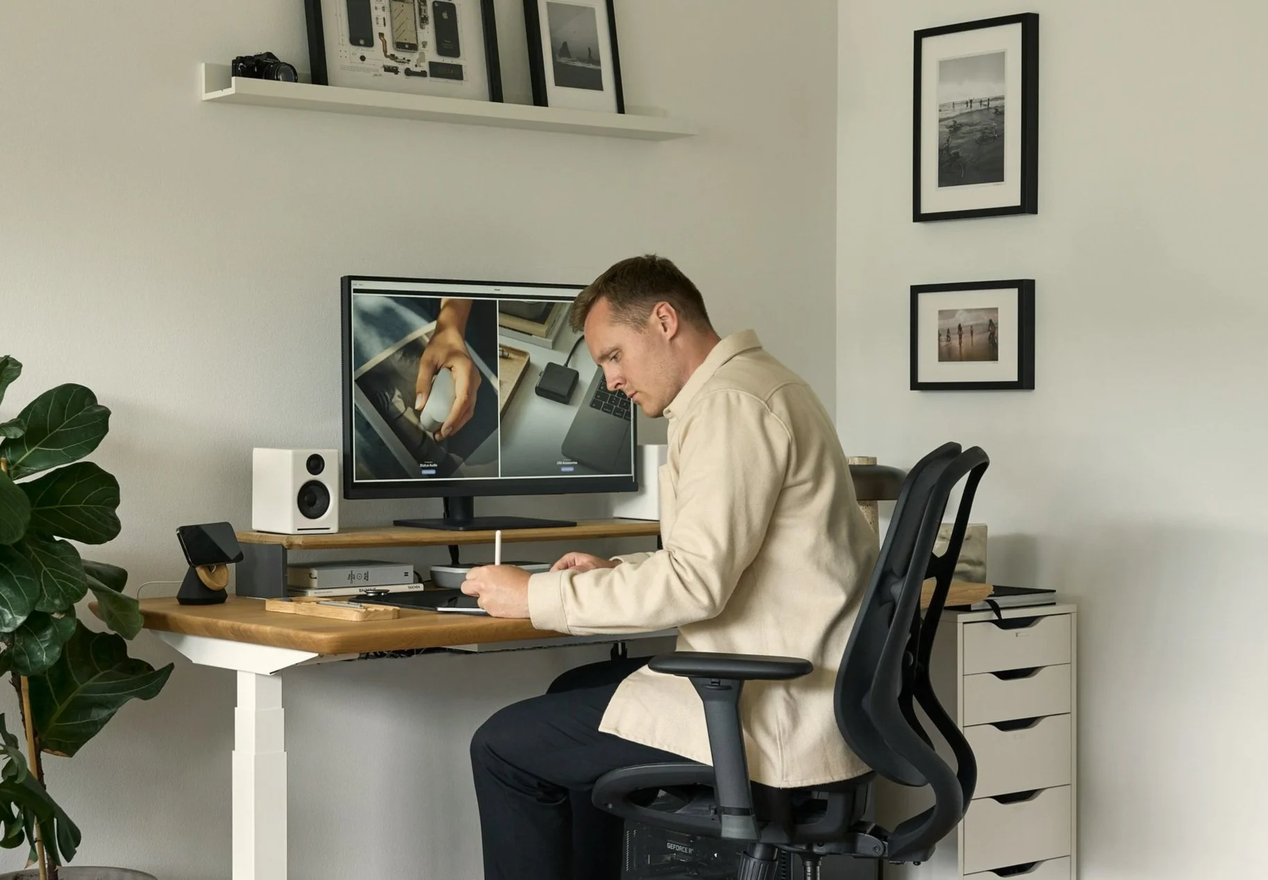 A man working at a wooden desk in a home office, with a dual monitor setup, a white speaker, and various electronic devices. He is sitting in a black ergonomic chair. The wall behind him features framed black-and-white photographs, and a large plant is visible on the left side.