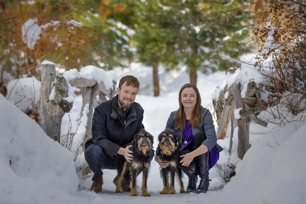 A man and woman kneeling on snow-covered ground with two dogs, outside in a winter landscape with snow, trees, and rustic wooden fences.