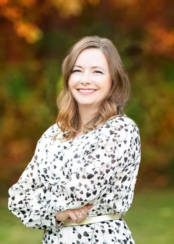 A woman with shoulder-length wavy hair smiling outdoors with autumn-colored trees in the background.