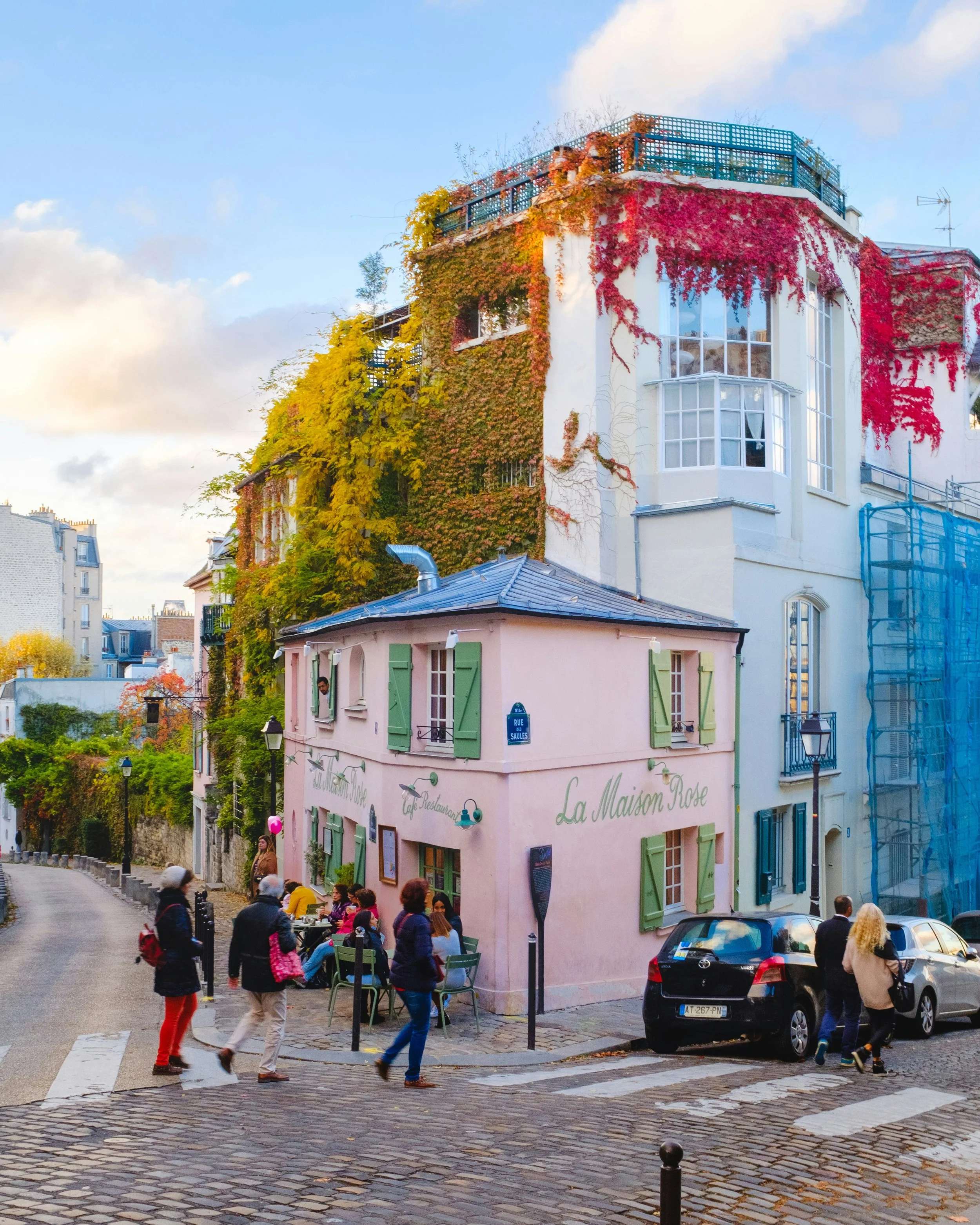 A street scene with a group of people walking across a crosswalk in front of a pink building with green shutters, with a sign that reads 'La Maison Rose'. The street is cobblestone, with cars parked along the side, and other buildings can be seen in the background. The weather is clear with some clouds.