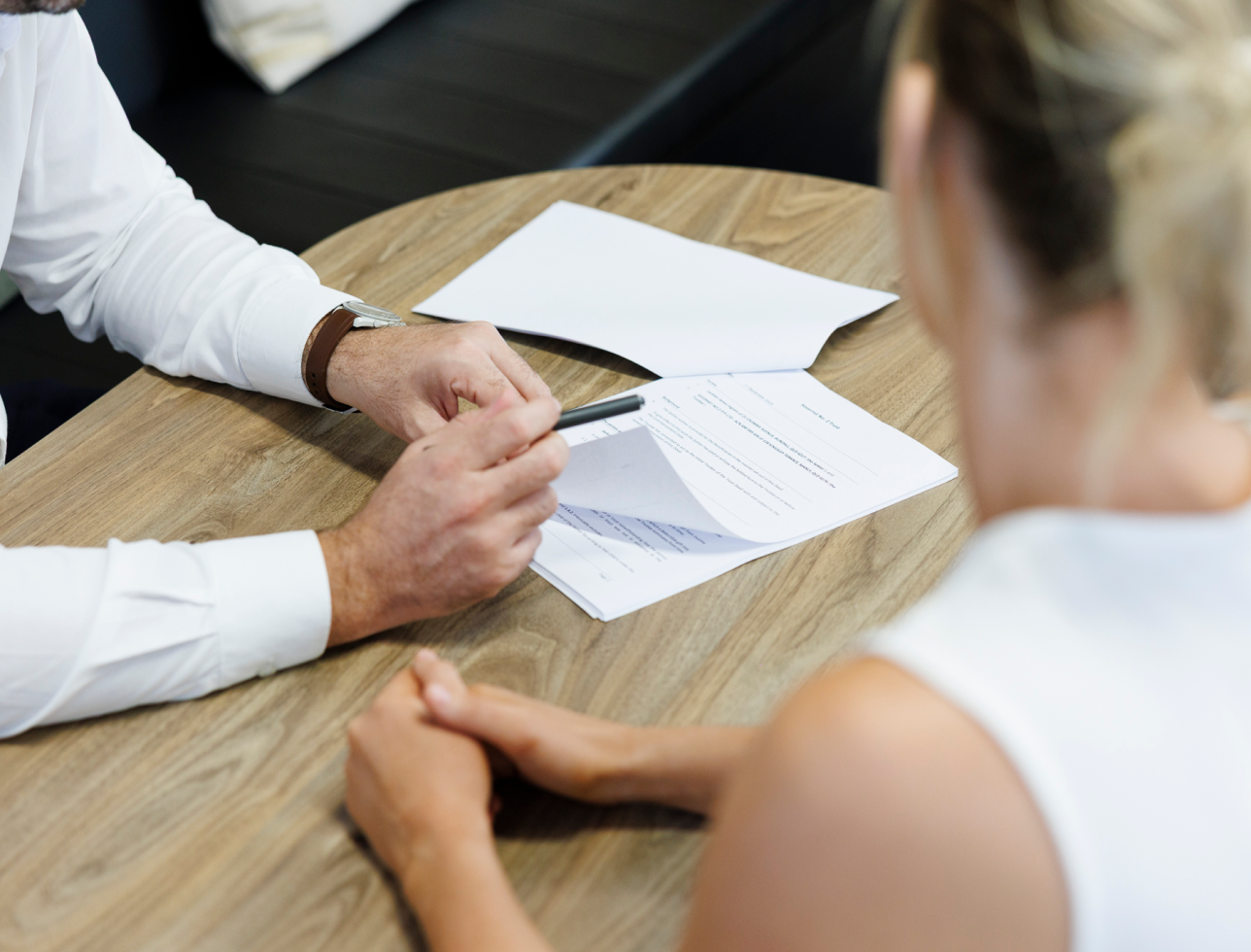 Two people sit at a wooden table during a job interview or business meeting, with documents and a pen on the table.