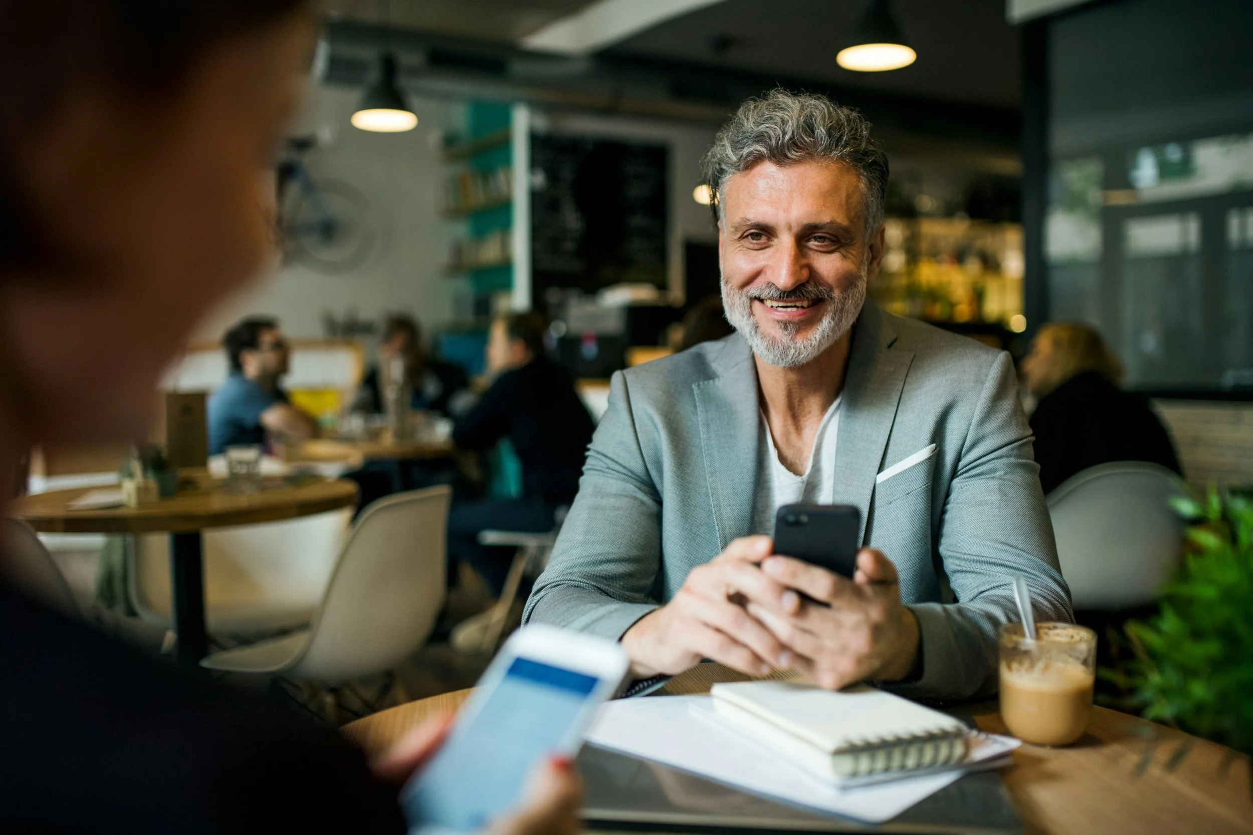 A smiling man with gray hair and a beard wearing a gray blazer, sitting at a table in a cafe or restaurant, engaging with a smartphone. In the background, there are other patrons and a modern interior.