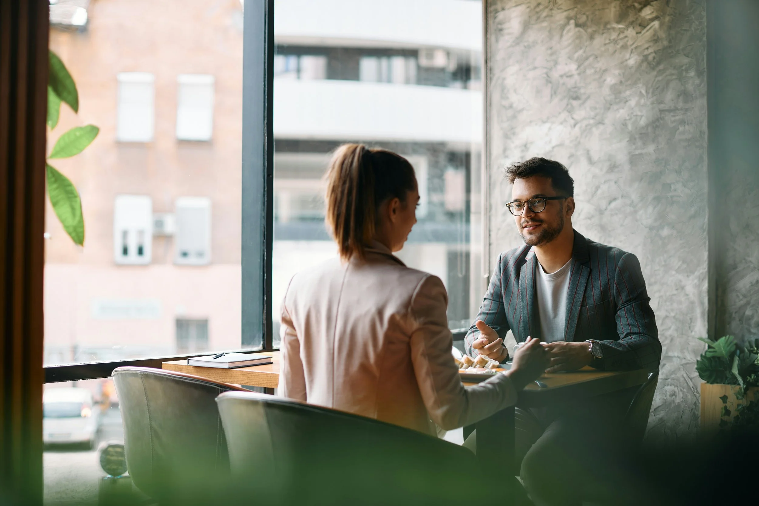 A man and woman sitting across from each other at a table near a window in a cafe, engaged in conversation. The man is wearing glasses and a striped blazer, while the woman has her hair tied back and is dressed in a blazer. Outside the window, buildings and a few cars are visible.