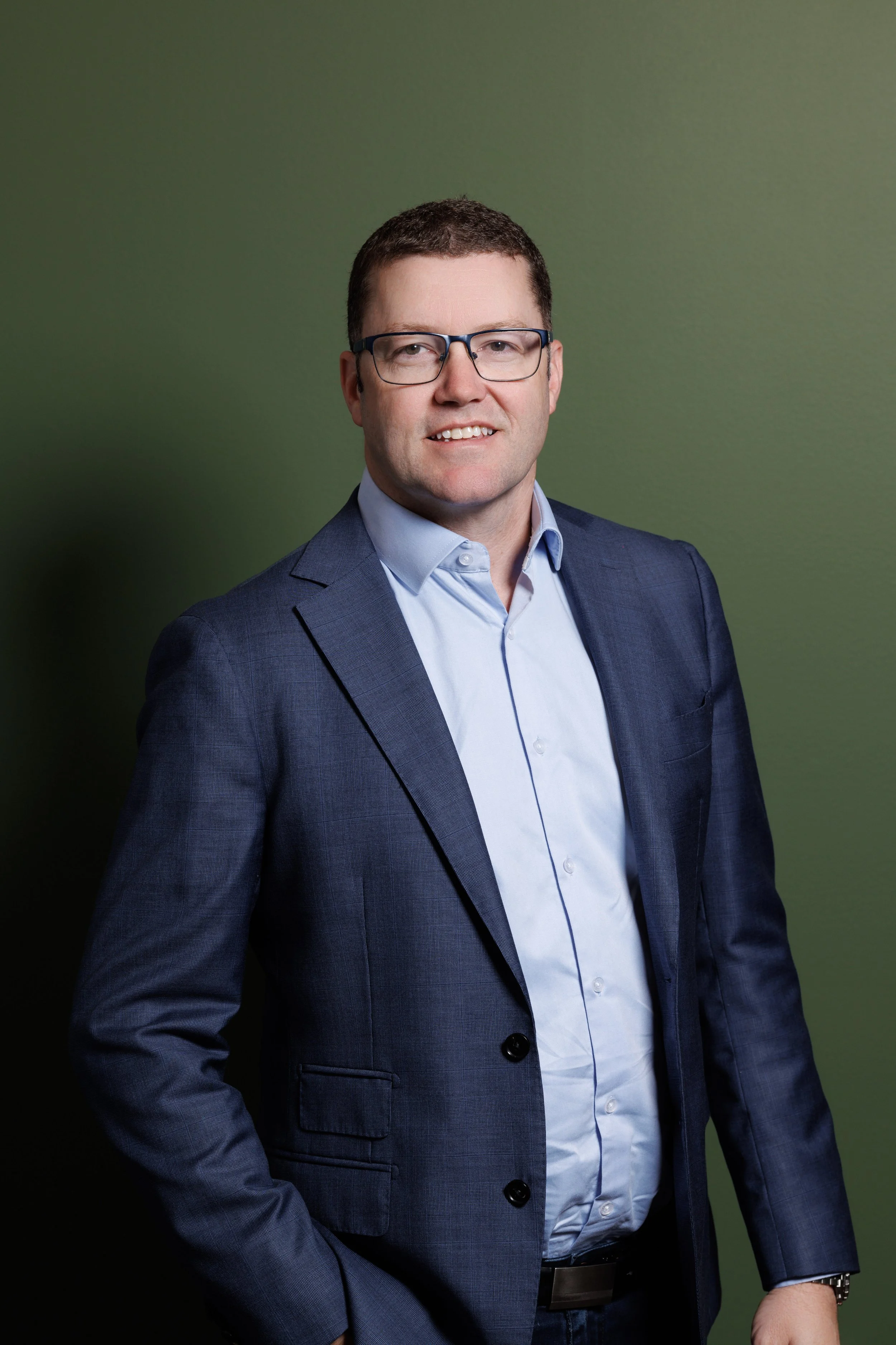 Headshot of a man in a navy suit and glasses, smiling, standing against a green background.
