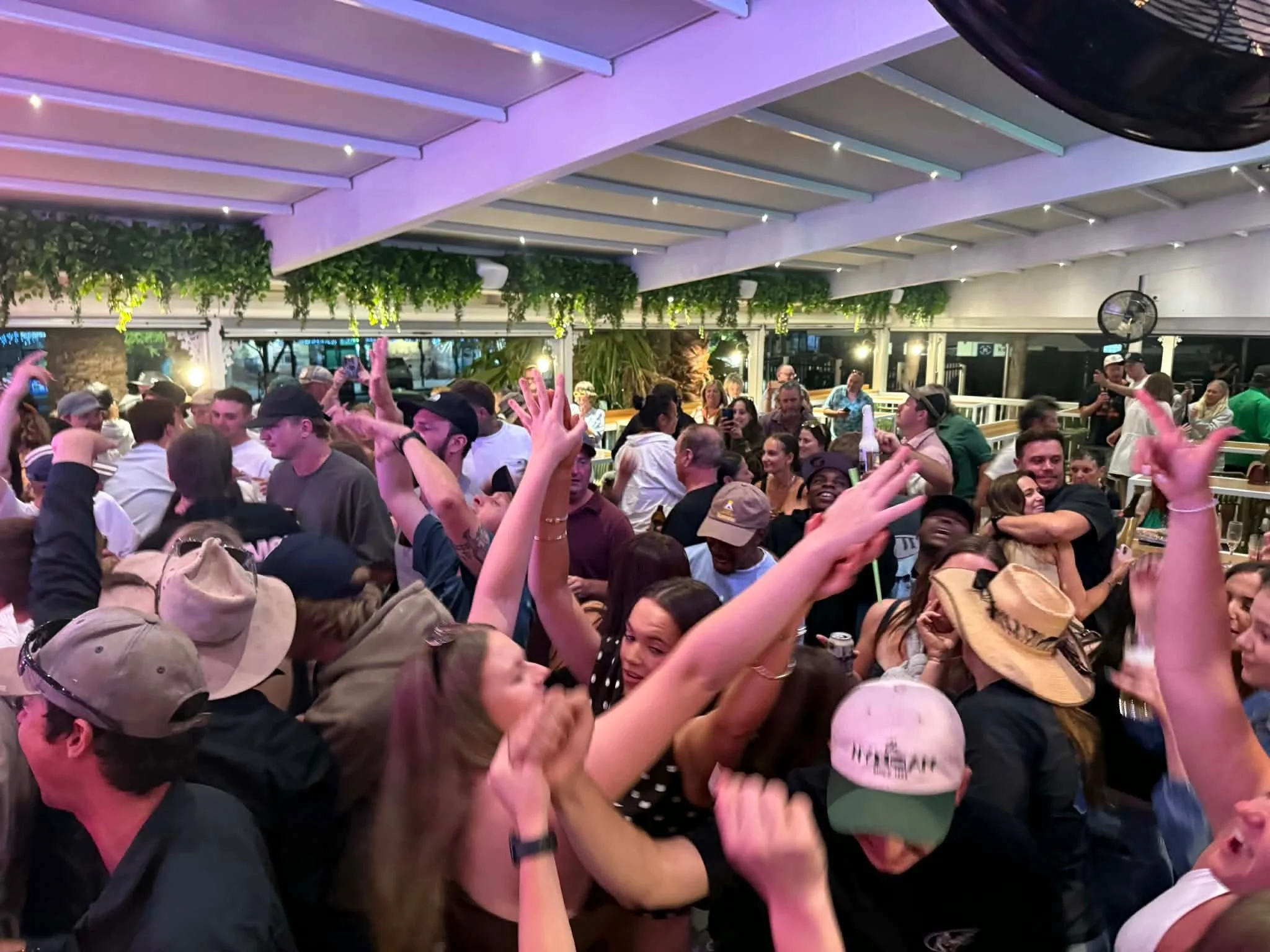 Crowd of people at an indoor event or party, some are wearing hats and holding drinks, with string lights and greenery hanging from the ceiling.
