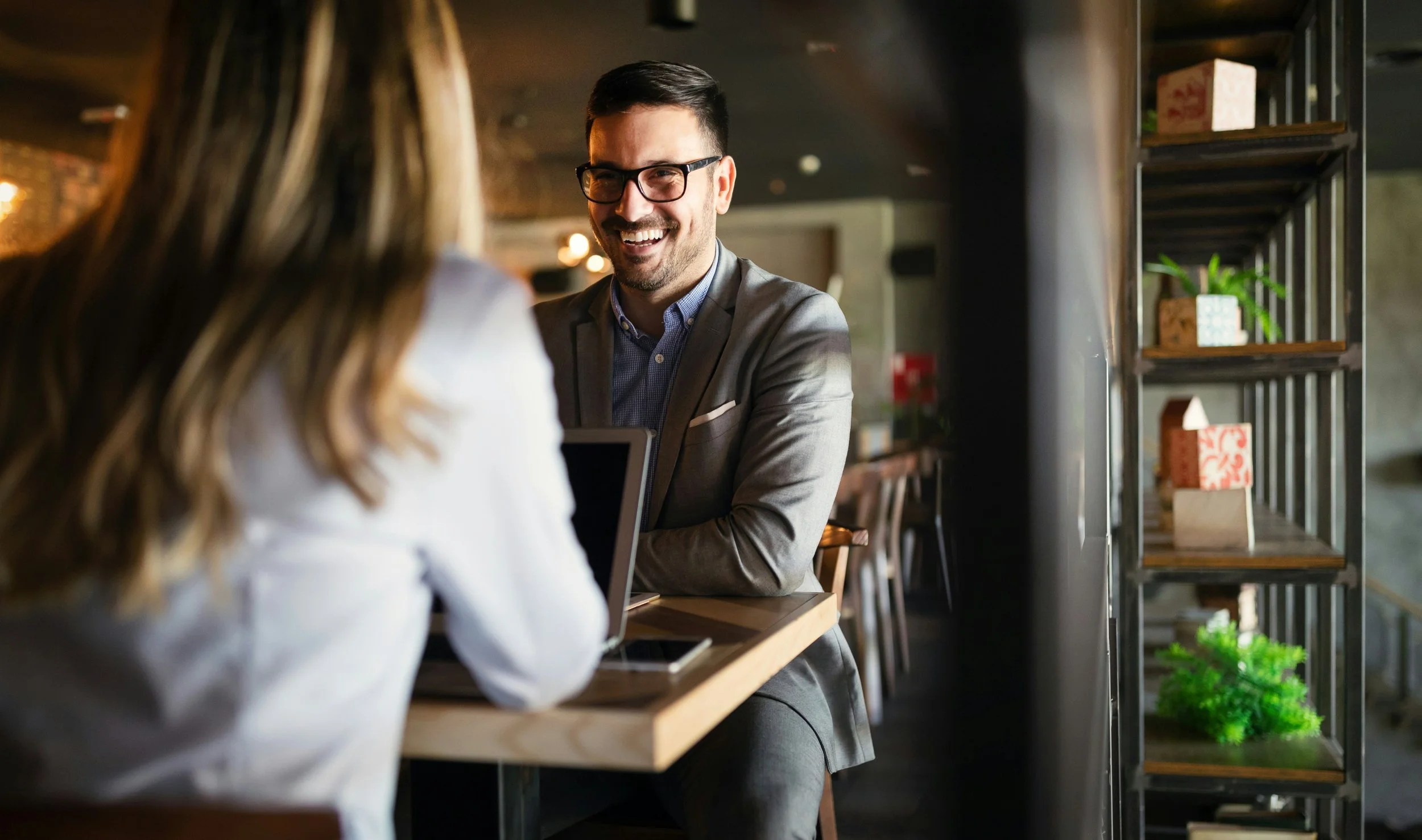 A man with dark hair, glasses, and a beard, wearing a gray suit, smiling and talking to a woman with long hair at a café or restaurant. The background includes wooden shelves with decorative items.