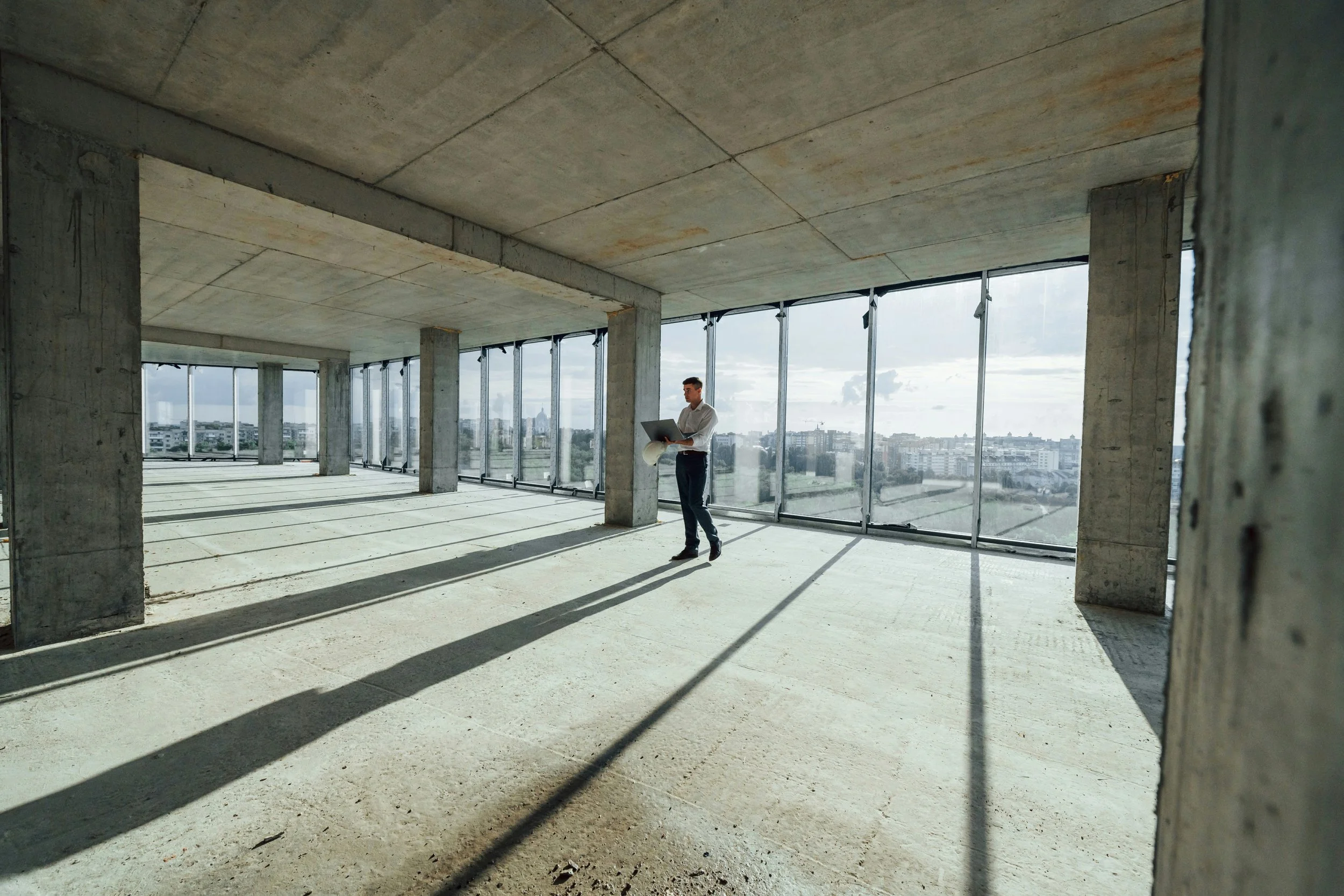 A man in formal attire holding a laptop inside an unfinished high-rise building with large windows and exposed concrete columns, overlooking a cityscape under a cloudy sky.