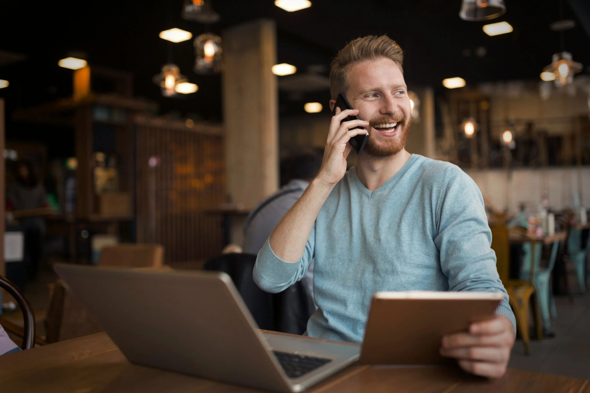 A young man with red hair and beard smiling while talking on his cellphone at a table in a cafe or restaurant.