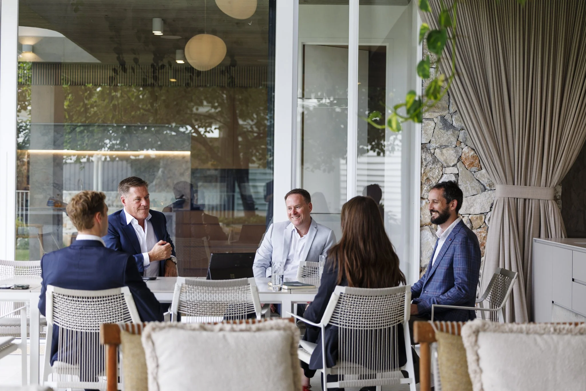A group of seven professionally dressed people, five men and two women, having a business meeting in a modern indoor space with large windows and stone wall accents.