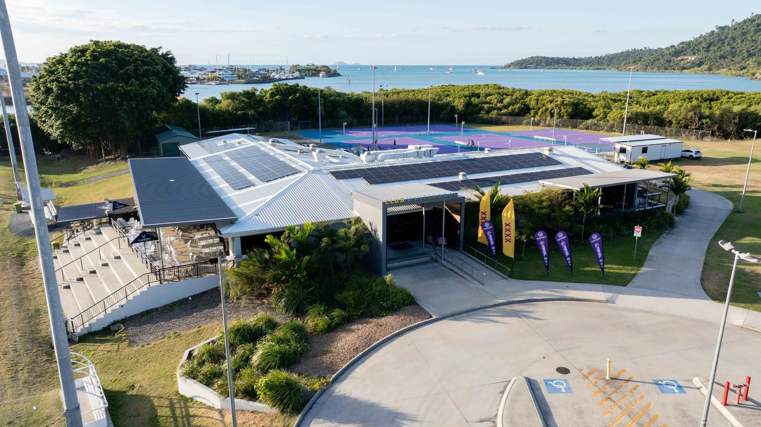 Aerial view of a community recreation area with tennis courts, a building with solar panels, and a scenic water background with boats and hills.
