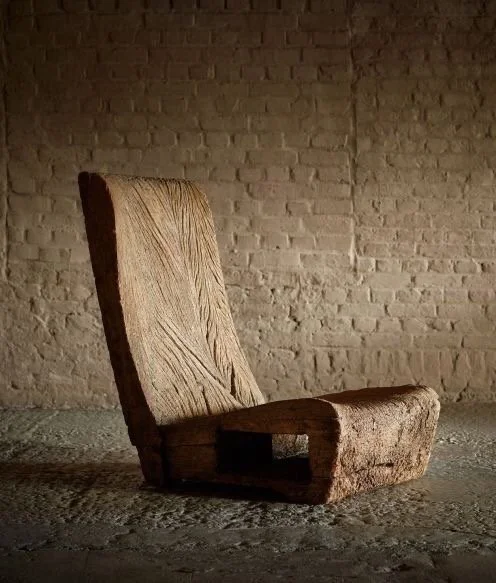A worn, beige upholstered chair with a high backrest and wide seat, placed against a beige brick wall in a dimly lit room.