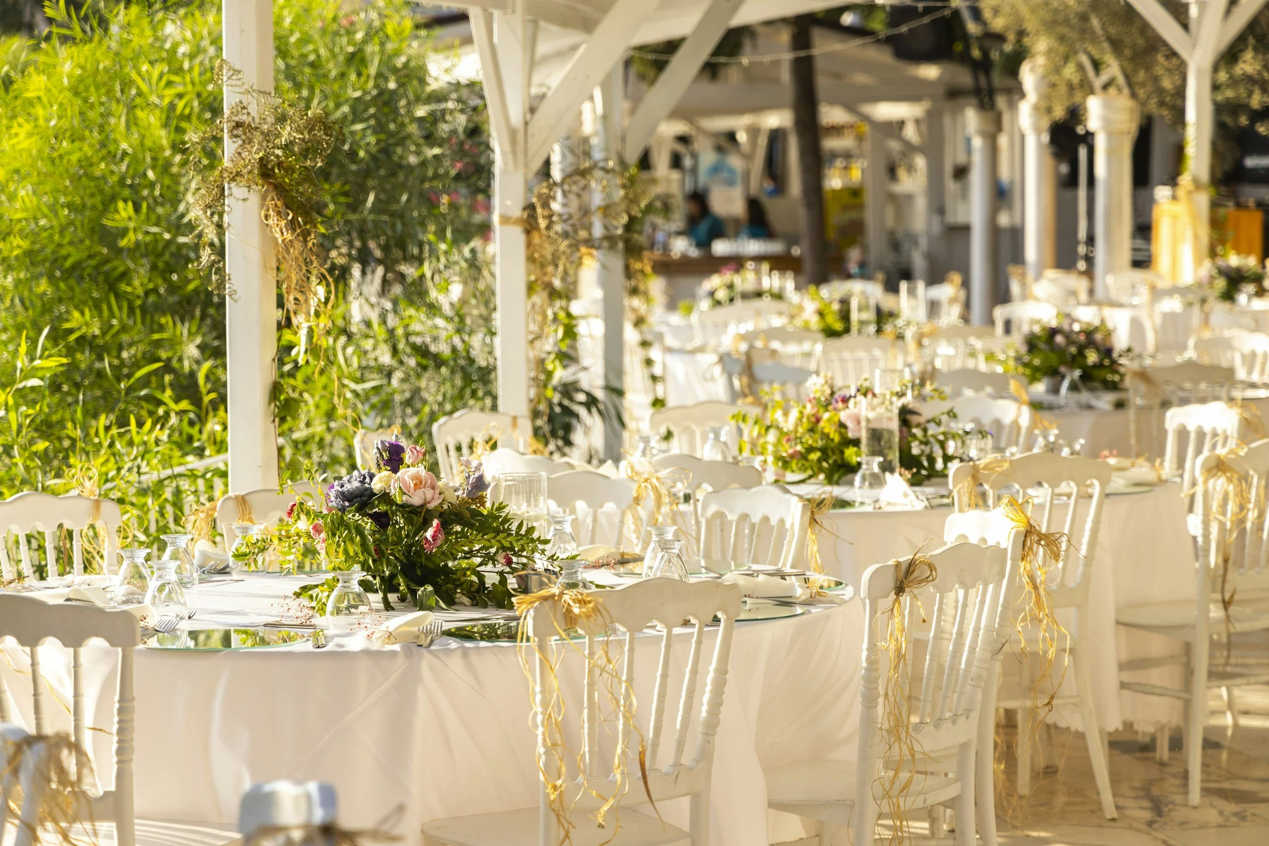 Outdoor event setup with round tables covered in white tablecloths, decorated with floral centerpieces, wine glasses, and golden tie-backs on white chairs.