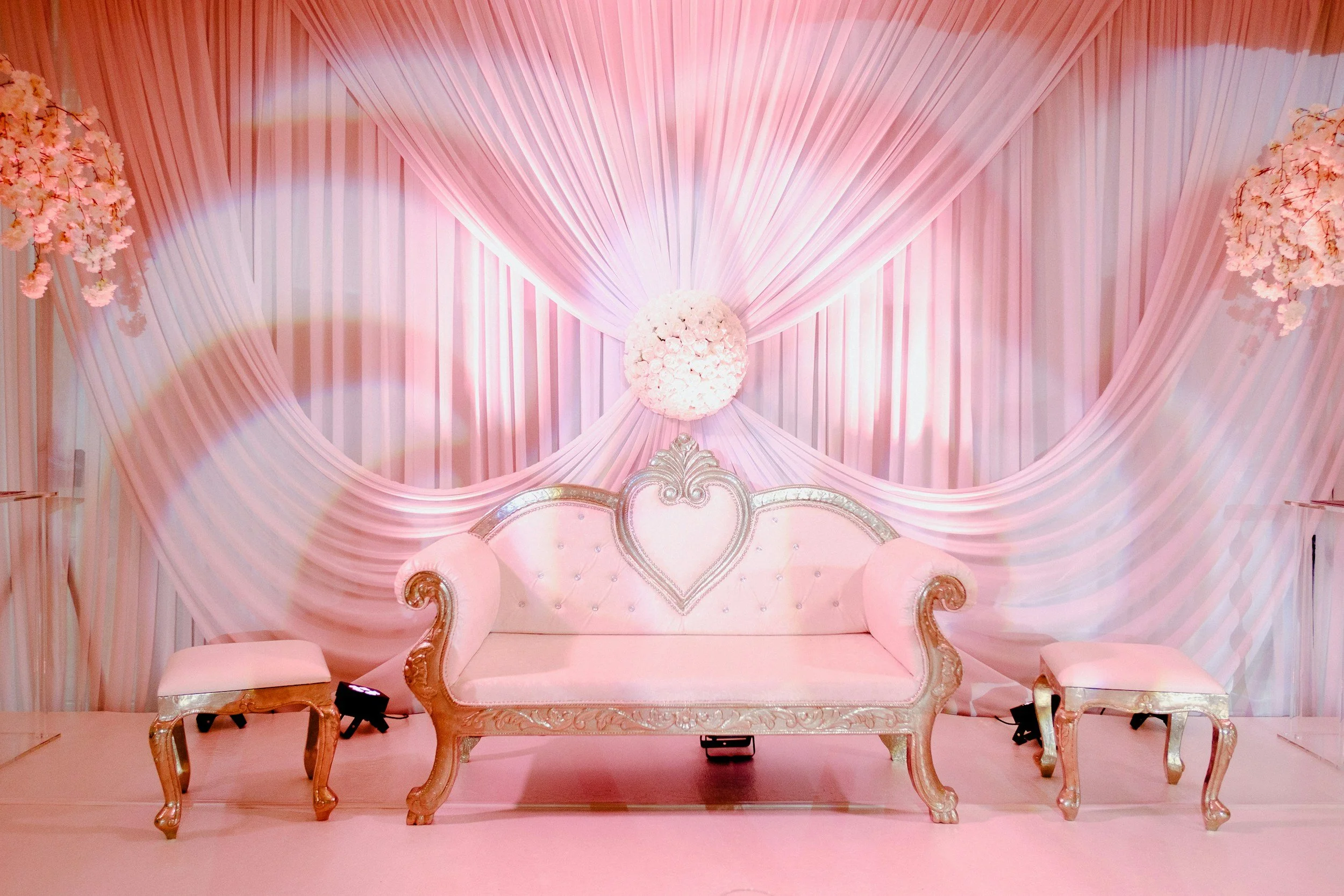 Elegant pink wedding chair with ornate gold trim and a heart-shaped tufted backrest, set against pink draped fabric backdrop adorned with floral arrangements.