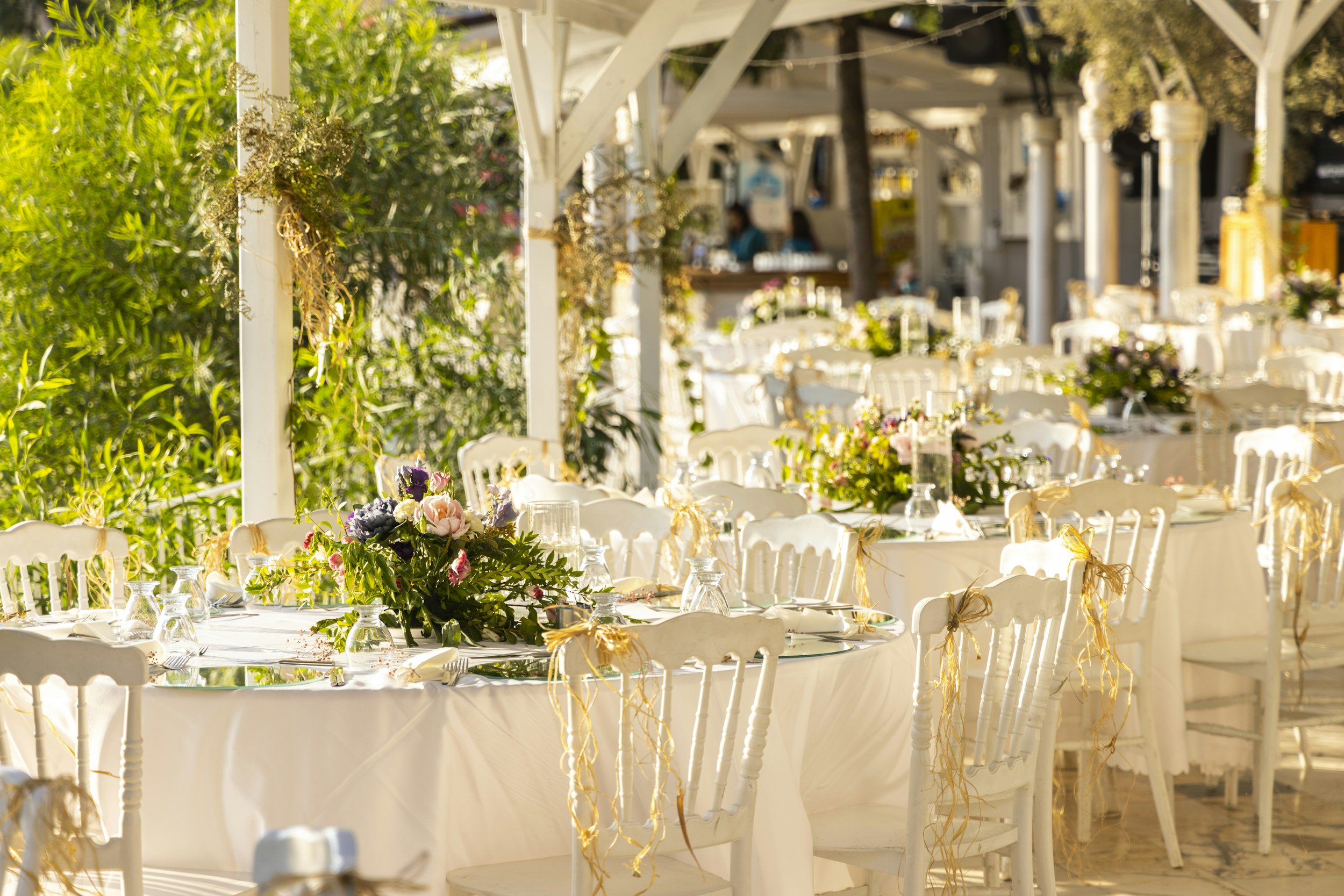 An outdoor banquet setup with long tables covered with white tablecloths and decorated with floral centerpieces. White chairs with ribbons are arranged around the tables, and the scene appears to be in a garden or patio with greenery and a white structure in the background.