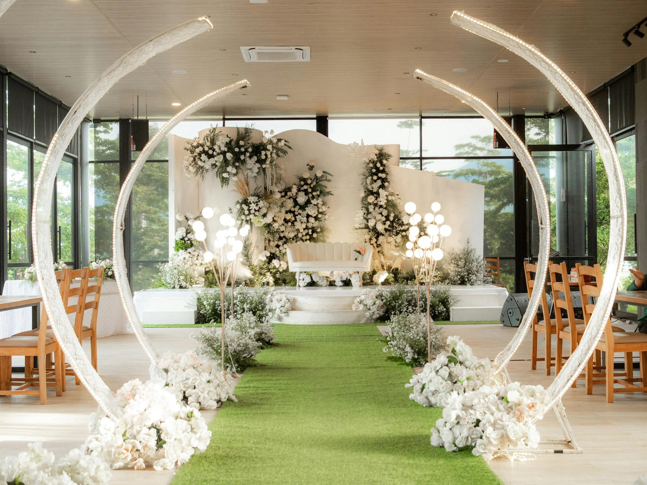 A wedding ceremony setup with an aisle covered in green turf and surrounded by white floral arrangements. There is a decorative backdrop with white flowers and greenery. The setting is indoors with large windows showing trees outside, and chairs are 