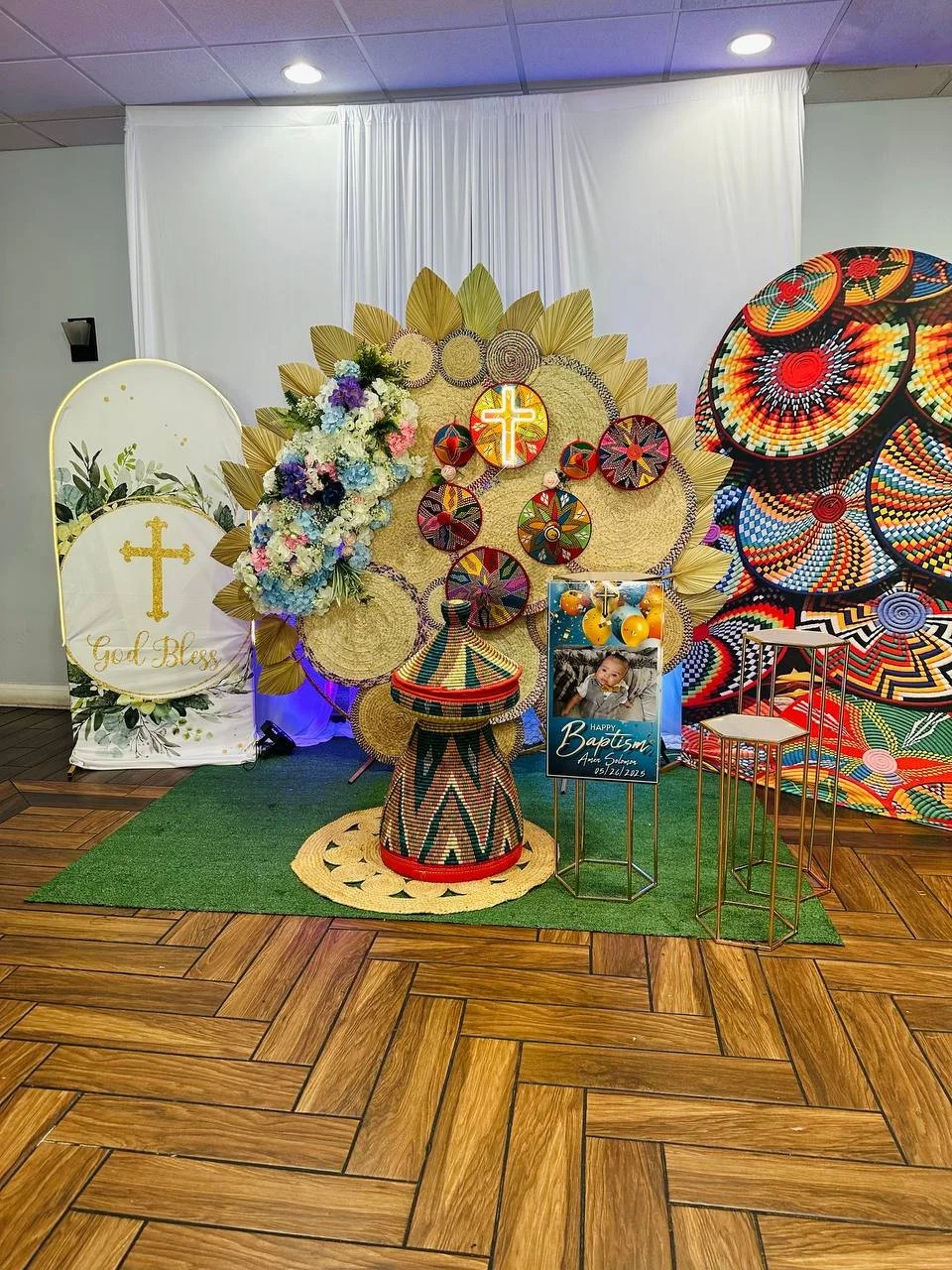 Decorative display for a baby's baptism, featuring floral arrangements, woven art, religious symbols, and a sign with a child's photo and baptism details.