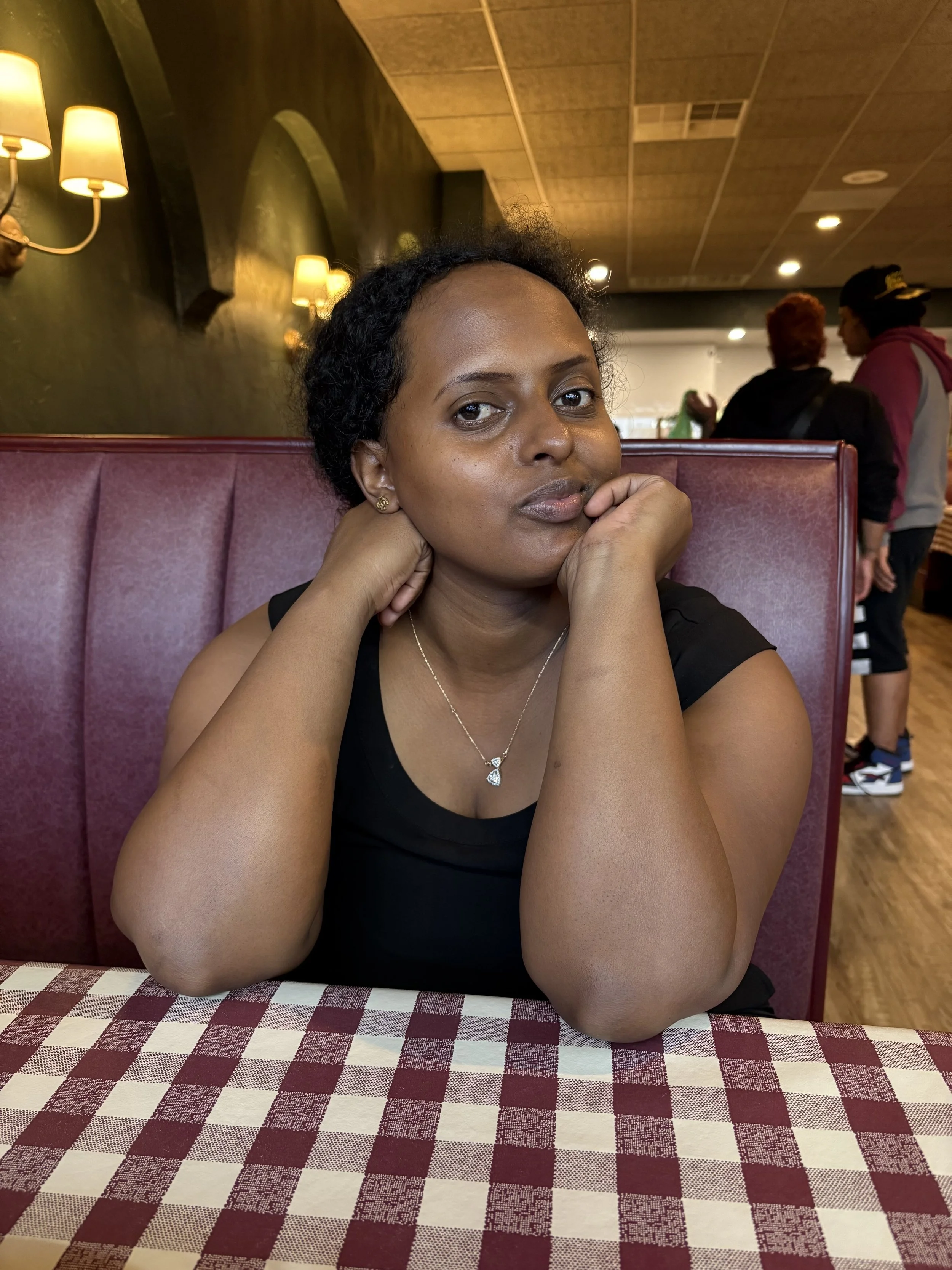 A woman with dark, curly hair sitting at a table with a red and white checkered tablecloth in a restaurant, resting her chin on her hands and looking at the camera.