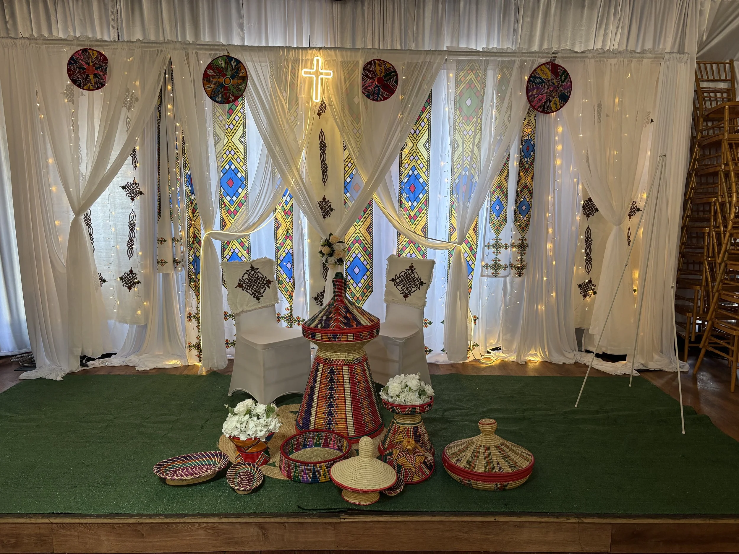 Decorative backdrop with white drapes, African patterned necklaces, and yellow lighted cross, with woven baskets and African-inspired items in front, set on a green turf floor.