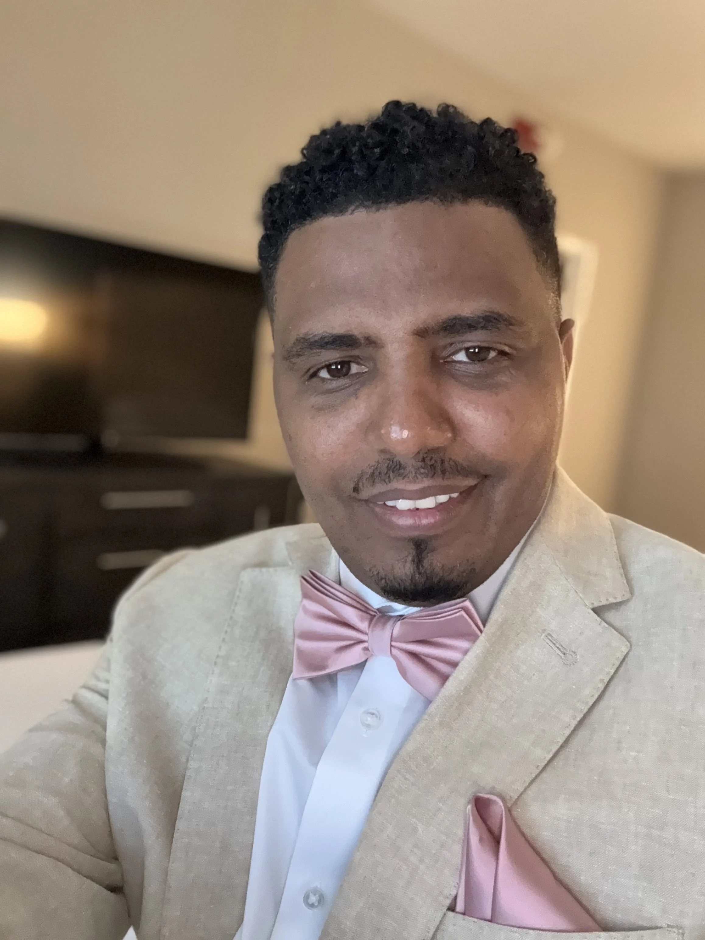 A man dressed in a beige suit, white shirt, and pink bow tie, smiling. His hair is styled in short, curly black hair. The background appears to be a room with a dark piece of furniture and a beige wall.