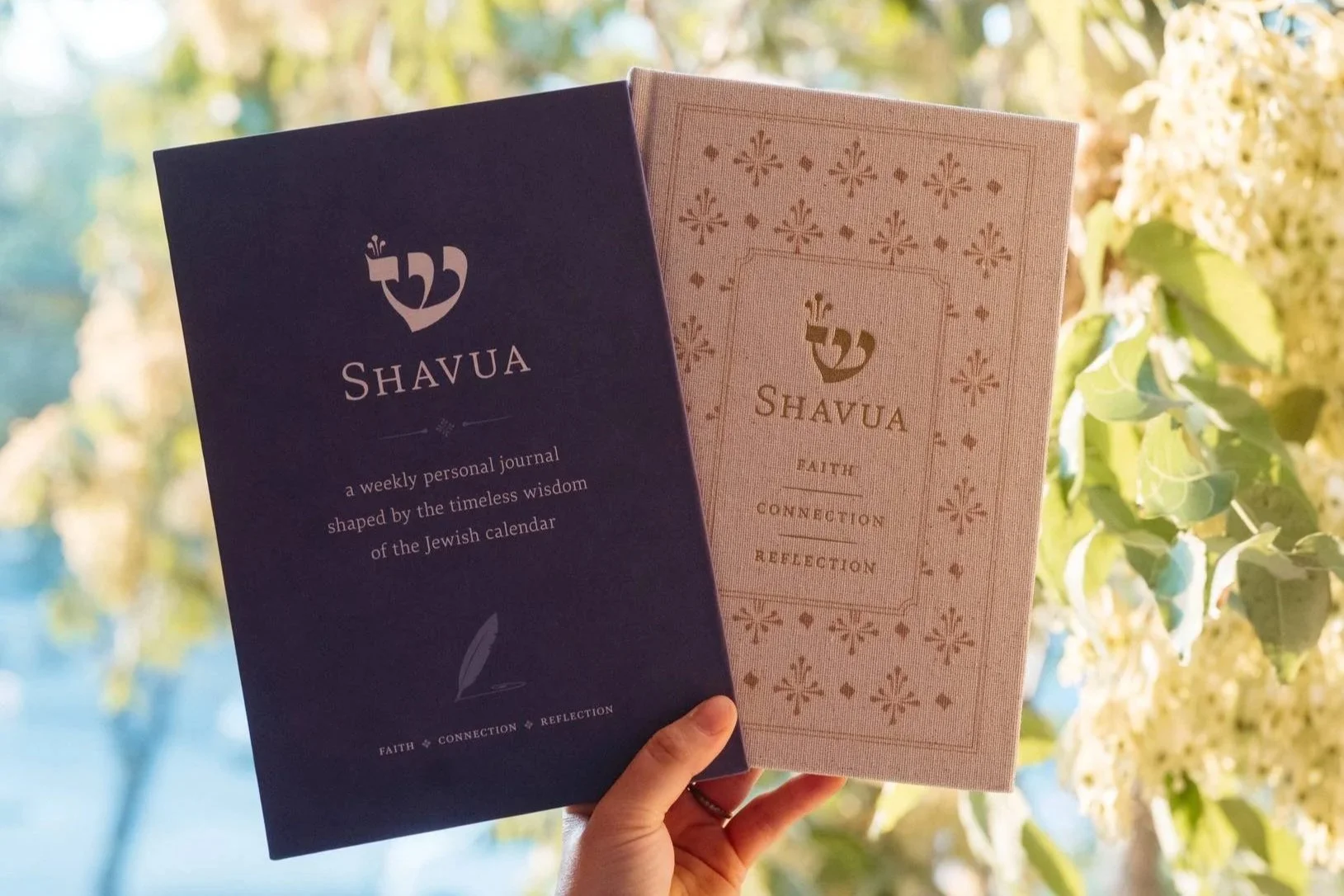 Two books titled 'Shavua', one in dark purple and the other in beige, held up against a background of green leaves and light-colored flowers. The dark purple book describes itself as a weekly personal journal shaped by the timeless wisdom of the Jewi