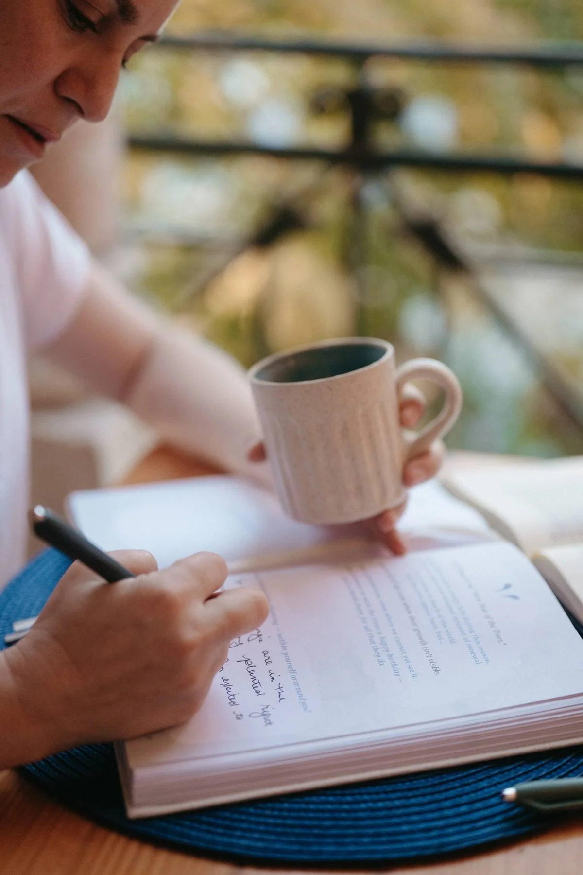 A person writing in a notebook with a pen, holding a ceramic mug, sitting at a table with a blue placemat, outdoors with a blurred background of trees and a railing.