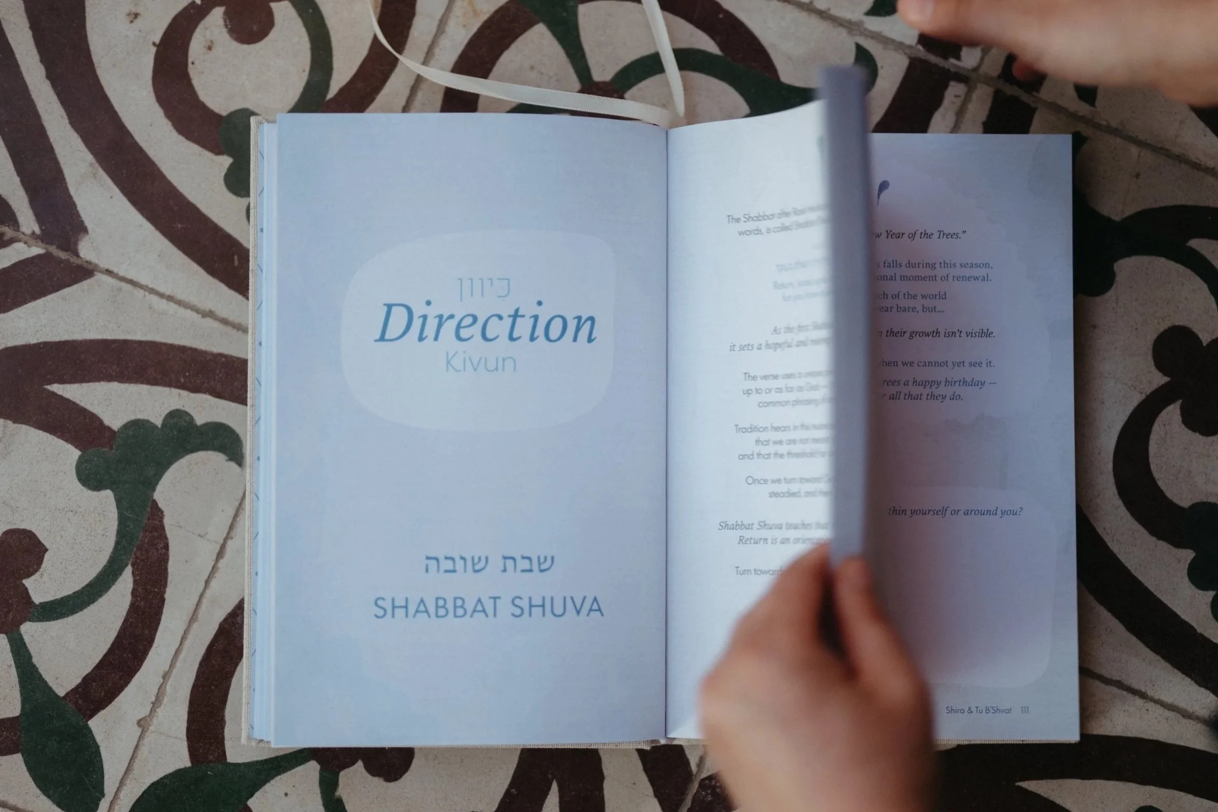 Open book on a tiled surface with a floral pattern, hands flipping the pages, with a focus on a page titled 'Direction' and 'Shabbat Shuva', written in English and Hebrew.