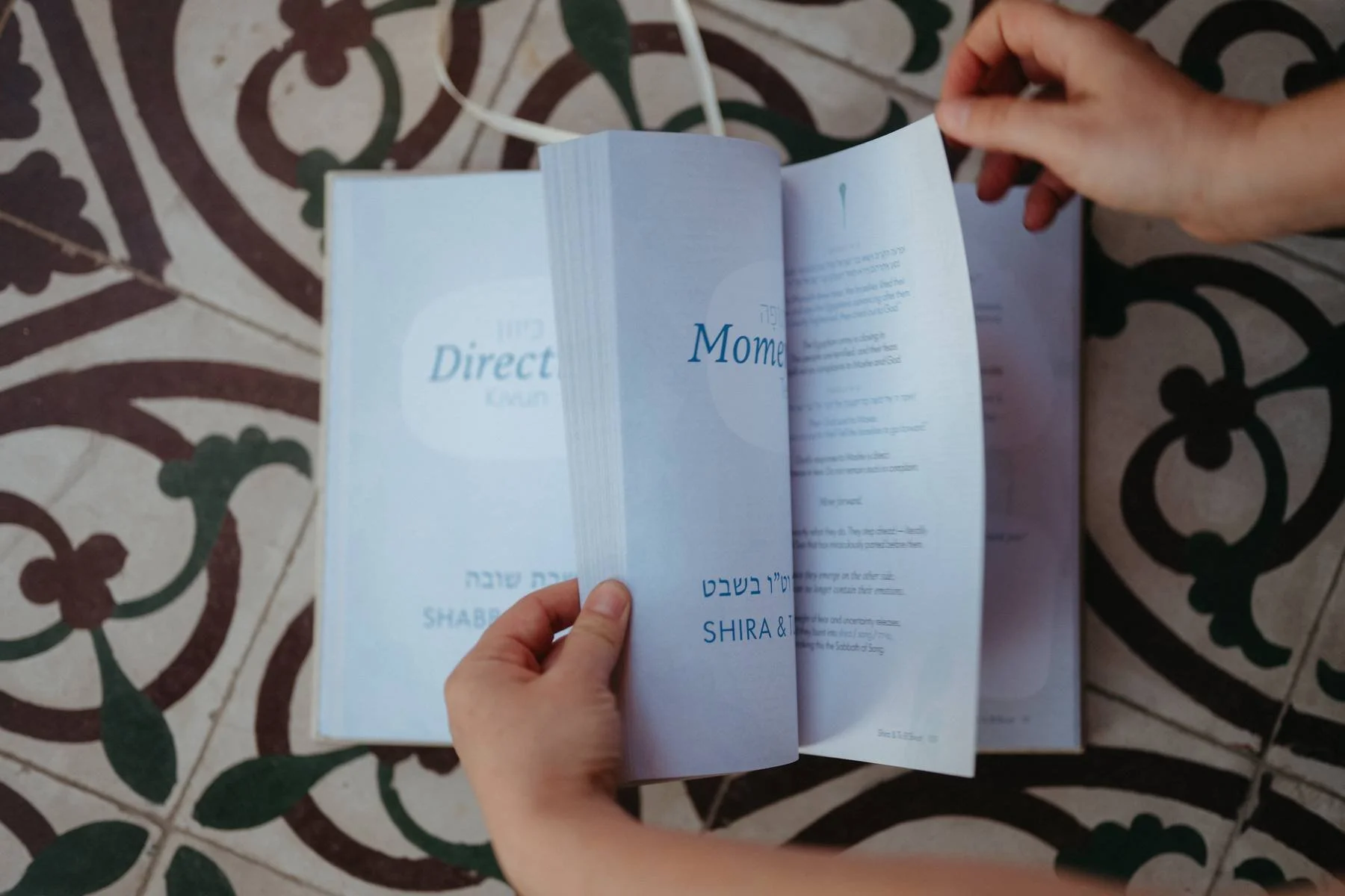 A hand flipping through a booklet or catalog on a decorative tiled floor with a floral pattern.