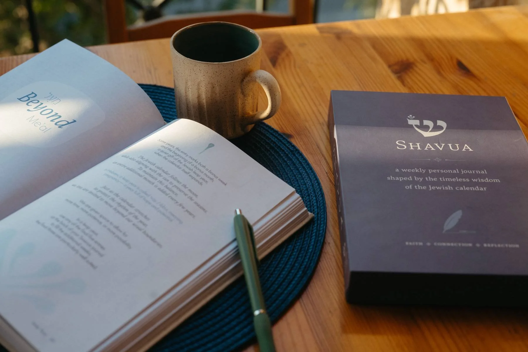 An open book, a ceramic mug, a pen, and a weekly personal journal titled 'Shavua' are placed on a wooden table with a blue placemat.